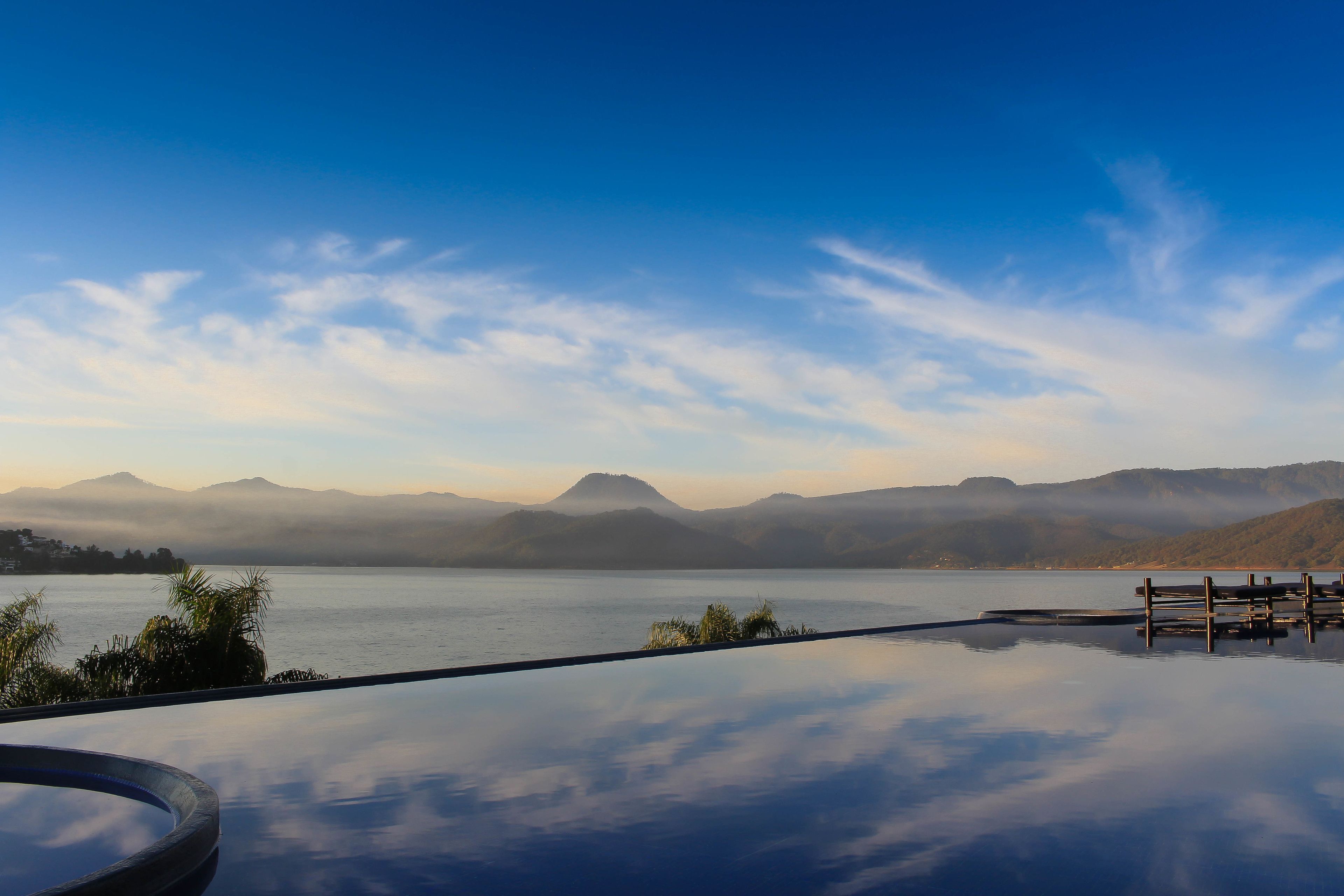 A serene lakeside view with a wooden dock extending into the water, surrounded by mountains and a clear sky.