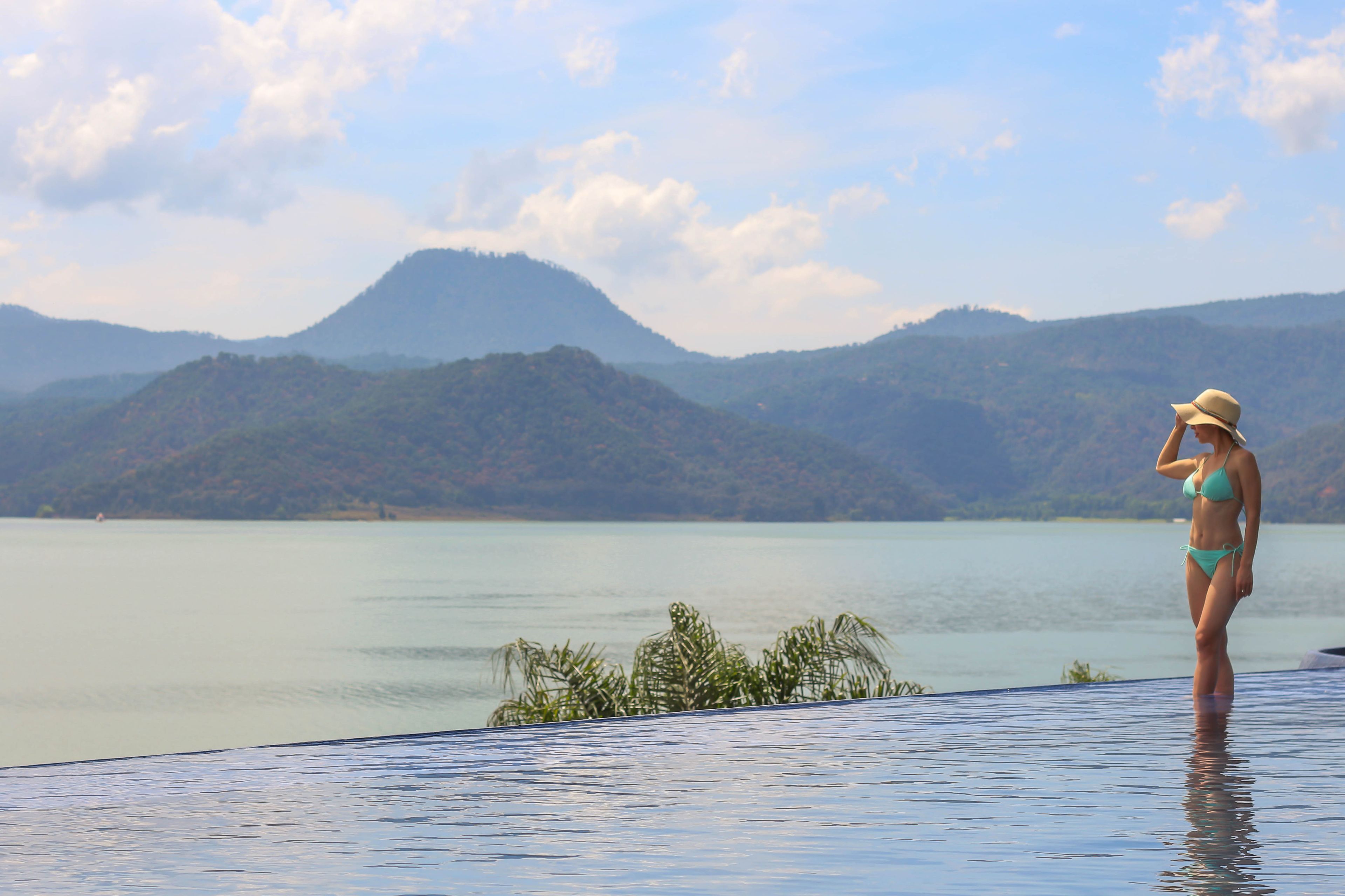 A person in a bikini standing by a pool with a mountainous landscape in the background.