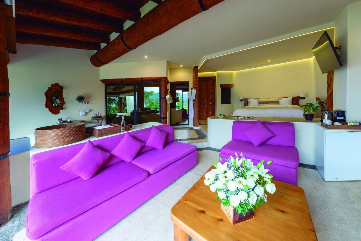 Ein modernes Hotelzimmer mit zwei violetten Sofas, einem hölzernen Couchtisch mit Blumenarrangements und einer Küchenzeile.