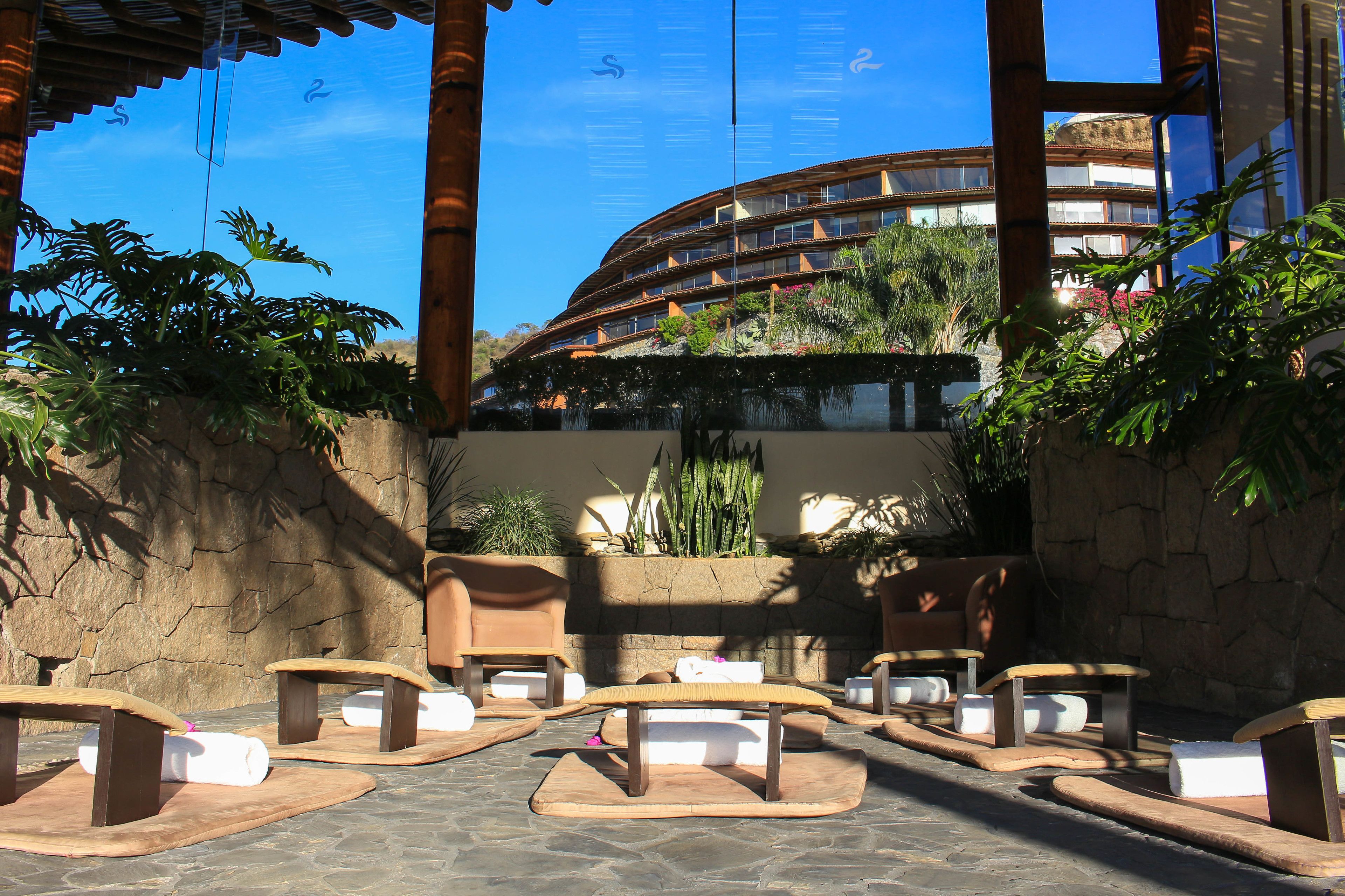 An outdoor seating area with wooden tables and benches, surrounded by plants and a stone wall.