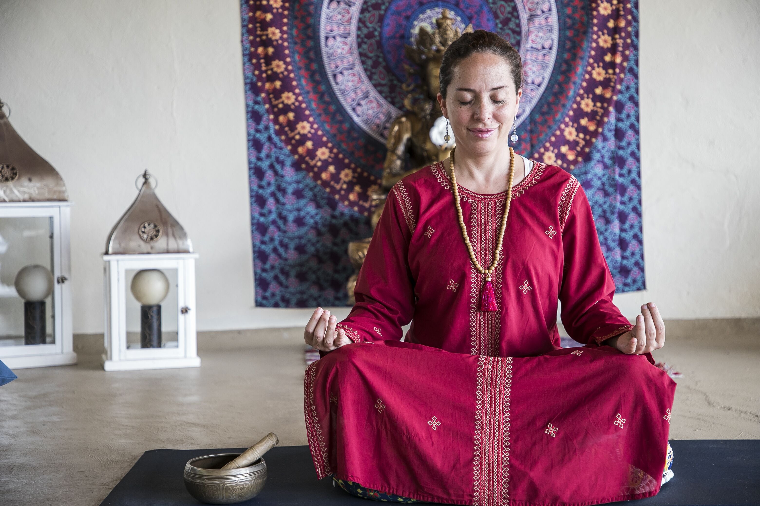 Una persona sentada en posición de meditación sobre una esterilla, con un tapiz decorativo y un pequeño altar de fondo.