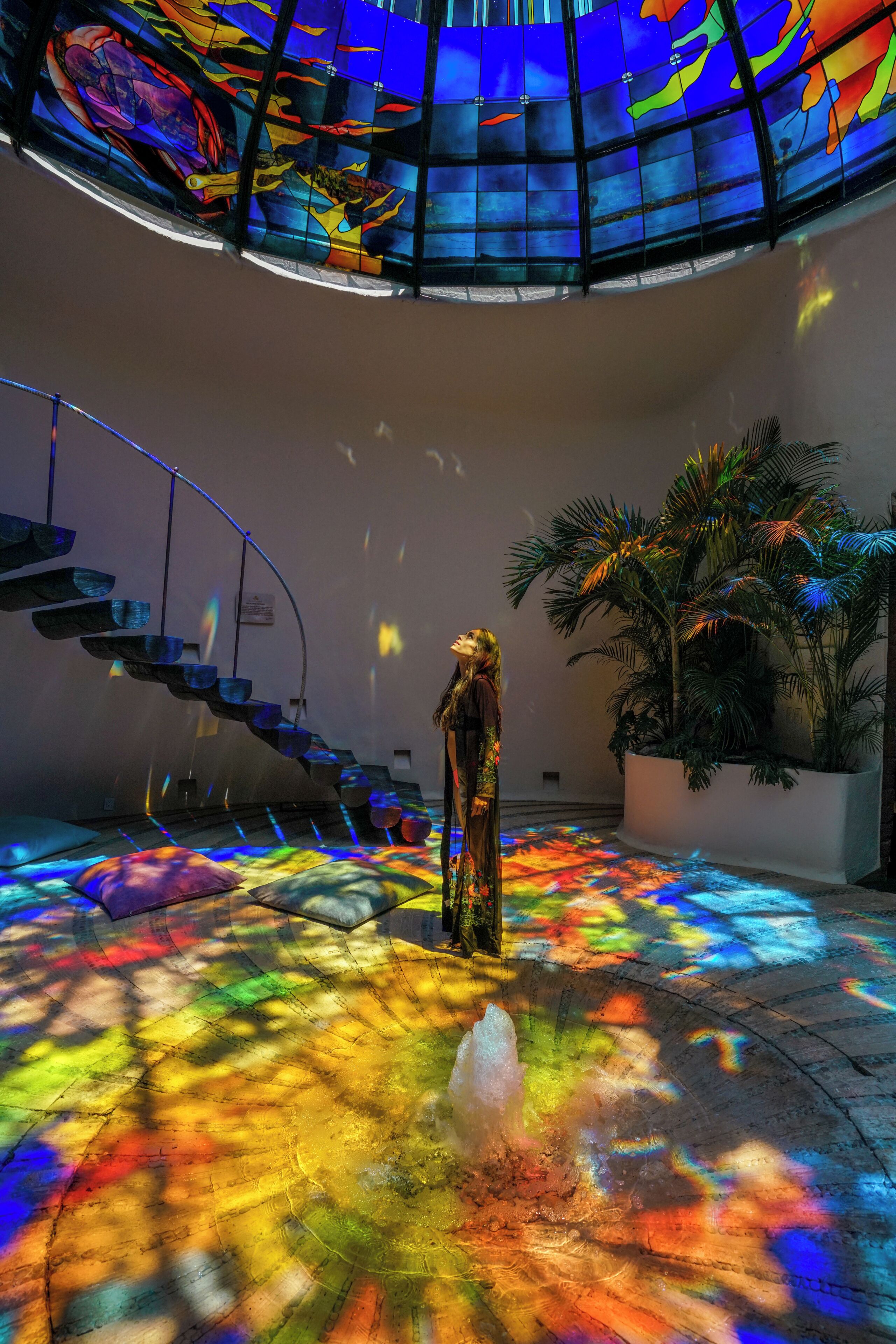 A person standing in a room with a colorful light installation on the floor, a staircase, and a large plant.