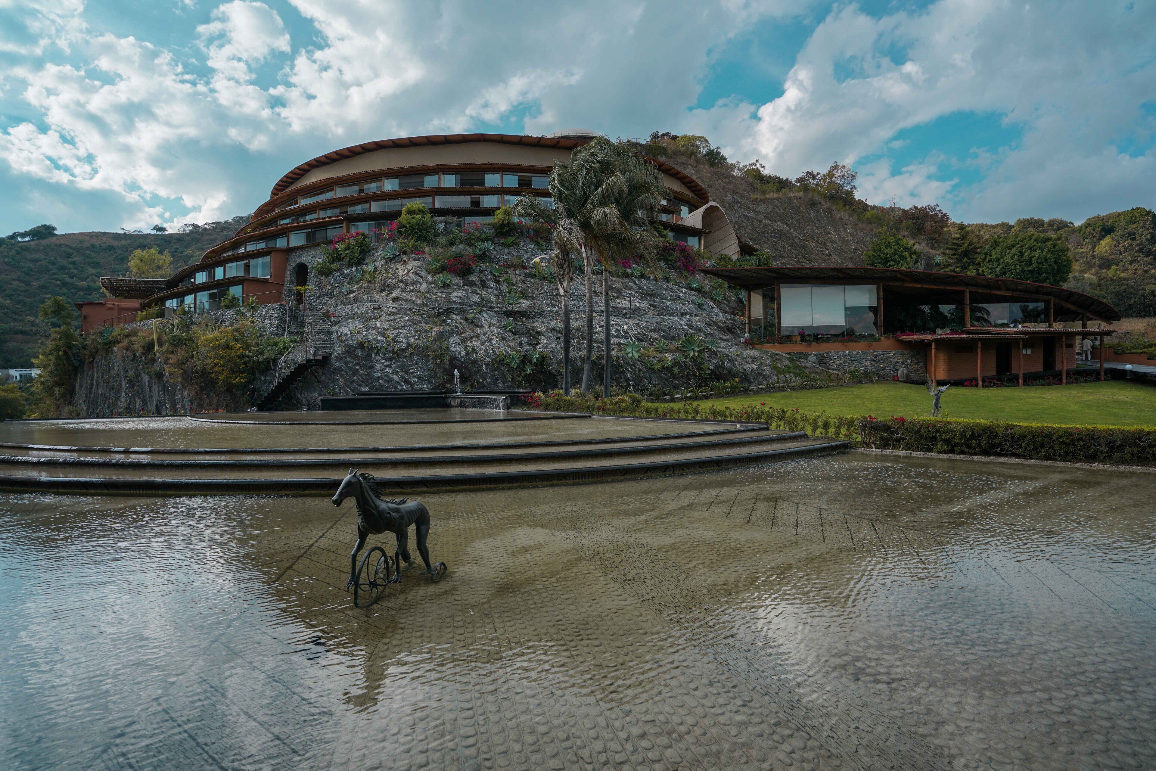 A large building with multiple floors, a fountain with a horse statue, and a landscaped area with a lawn and trees.