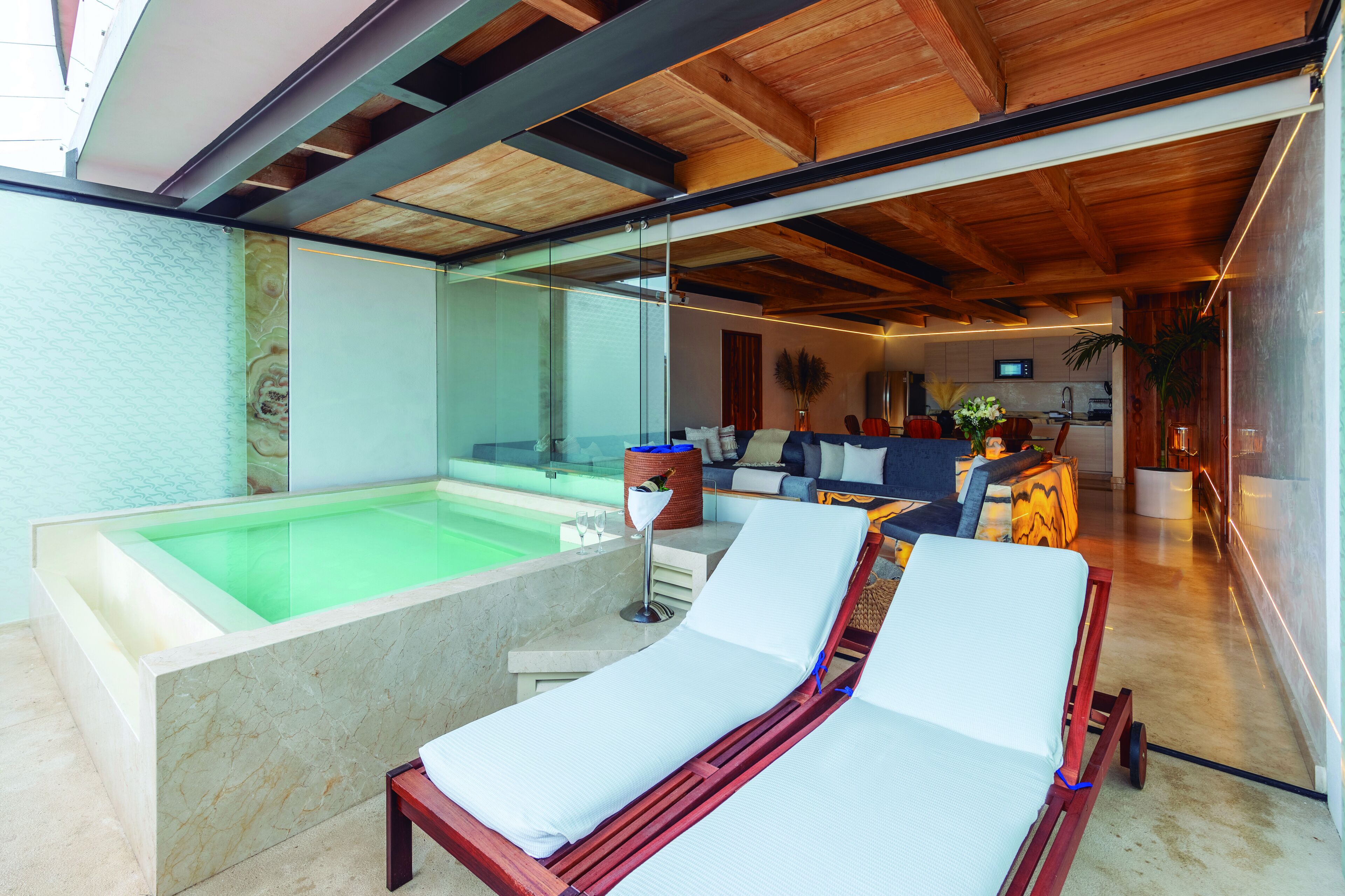 A modern indoor pool area with lounge chairs, a seating area with a sofa and coffee table, and a kitchen in the background.