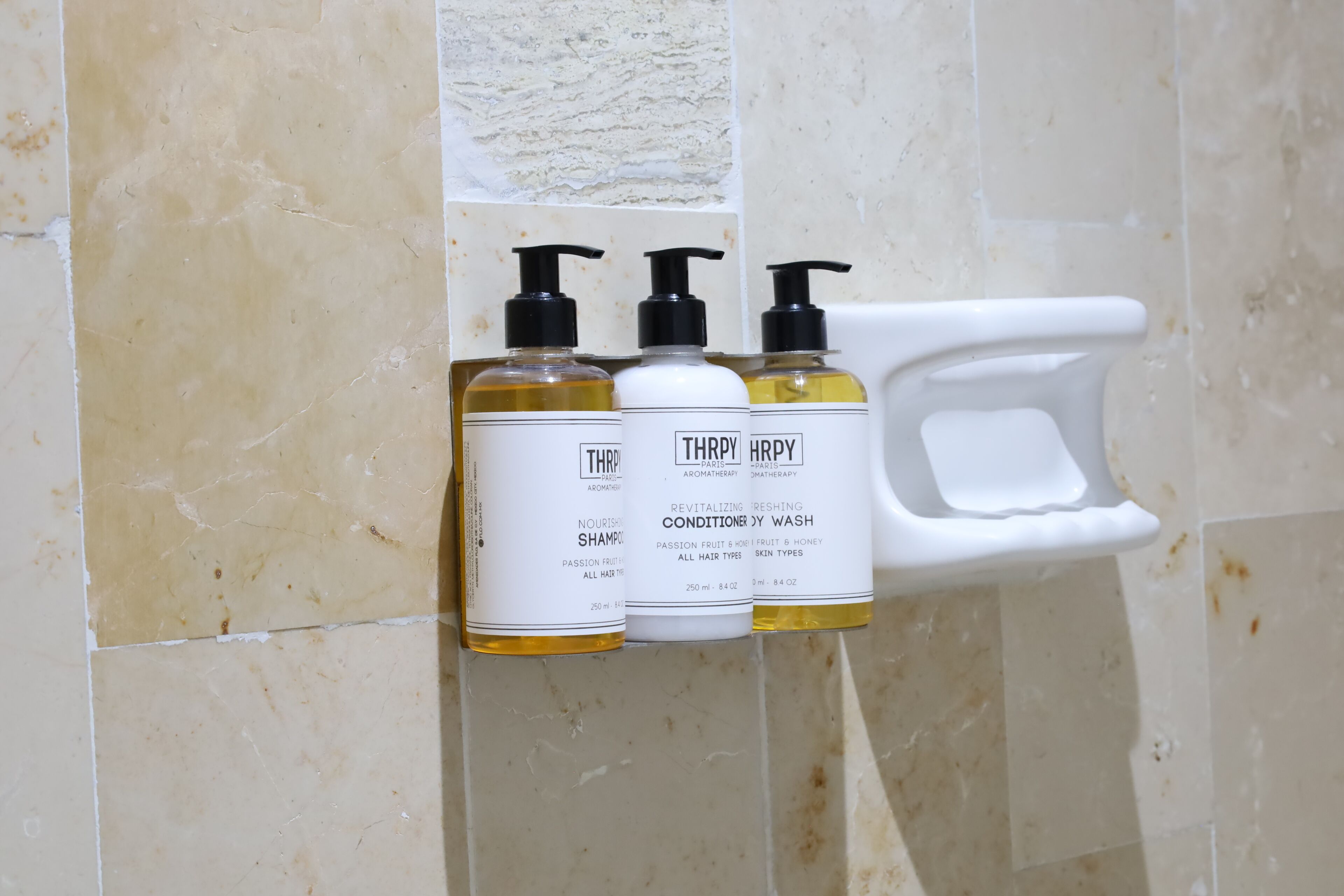 Tres botellas de Champú y Acondicionador Nutritivo Thrifty sobre un estante blanco frente a una pared de azulejos.