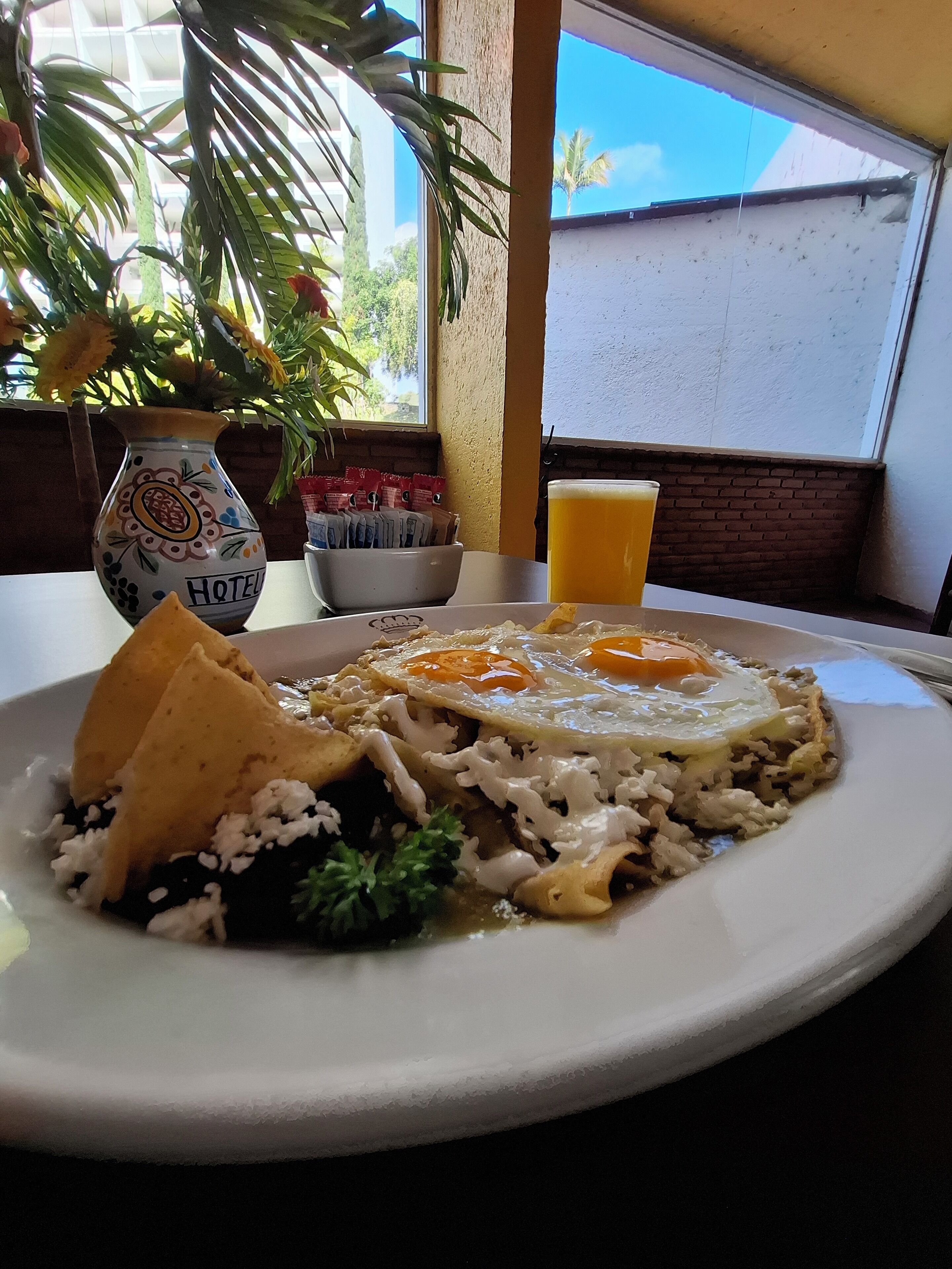 A plate of food with rice, fried eggs, and a tortilla, a glass of orange juice, and a decorative vase with a plant.