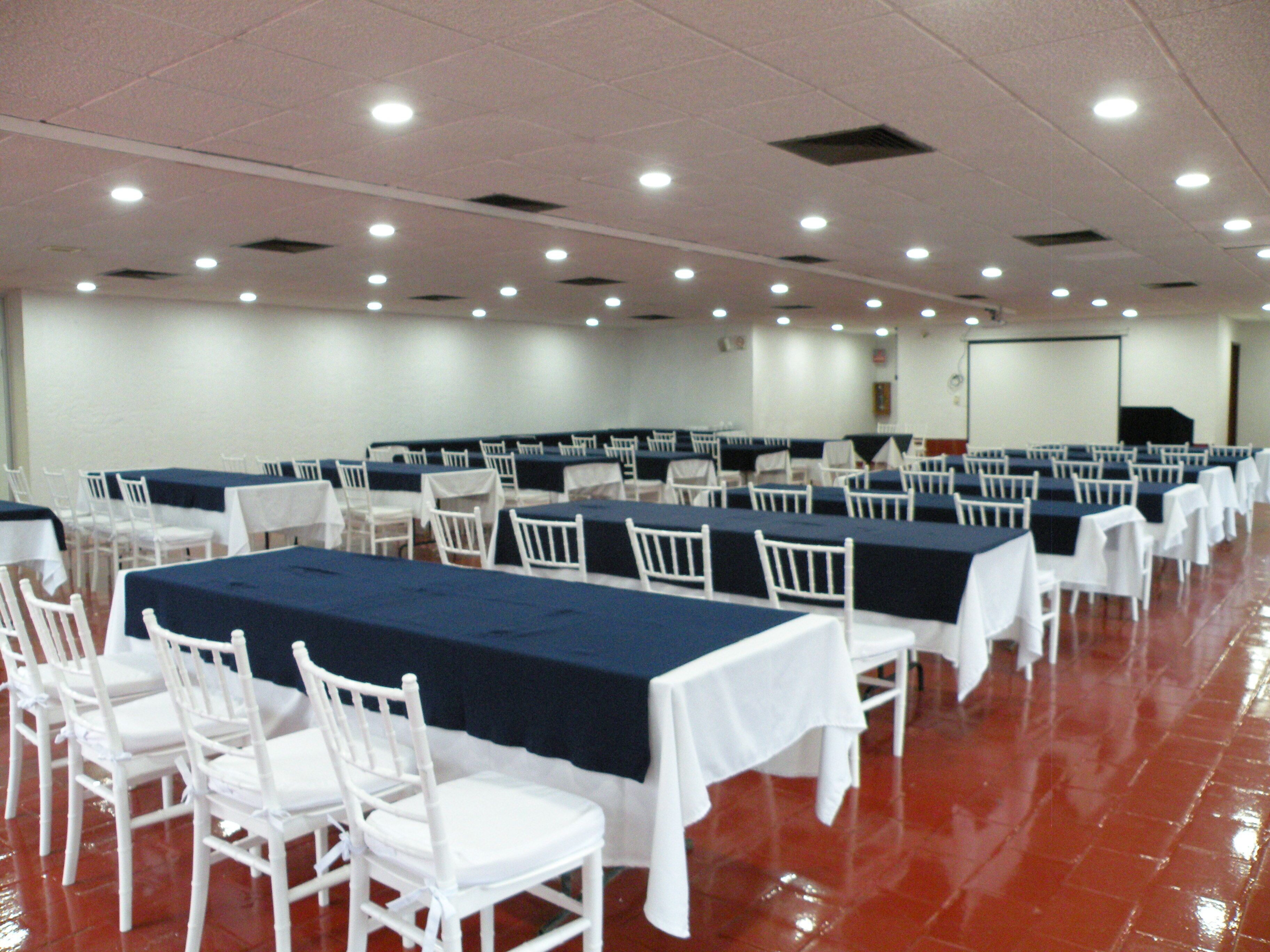 Un salón de conferencias con filas de mesas y sillas, una pantalla para proyector y una pizarra.