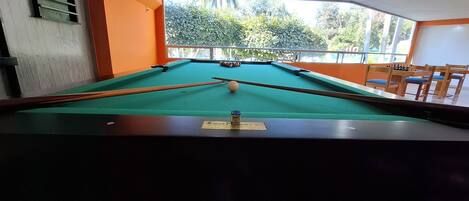 Una mesa de billar con superficie de fieltro verde, rodeada de paredes anaranjadas y una ventana que da a vistas de follaje.