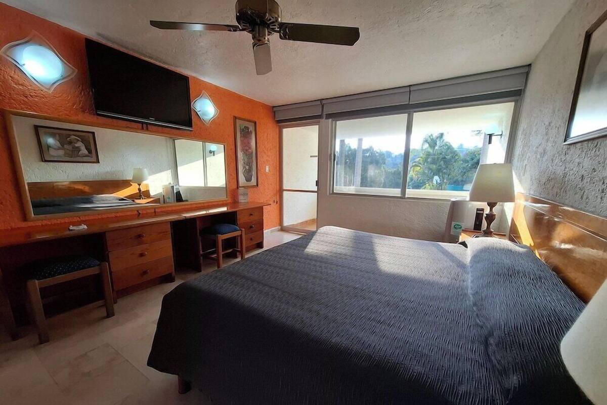 Un dormitorio con una cama grande, un escritorio, una silla, un televisor y una ventana con vista a árboles.