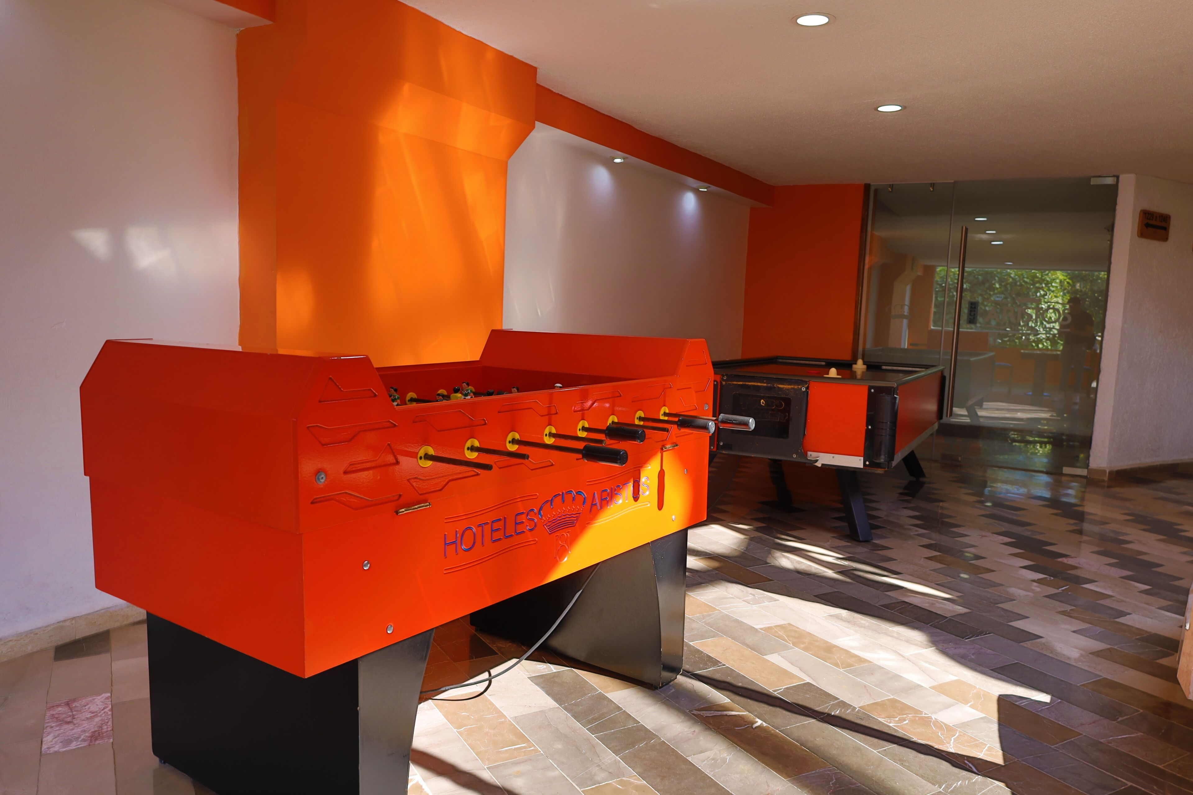 Una mesa de fútbol de color rojo en el vestíbulo de un hotel, con piso a cuadros y puerta de cristal que da al exterior.