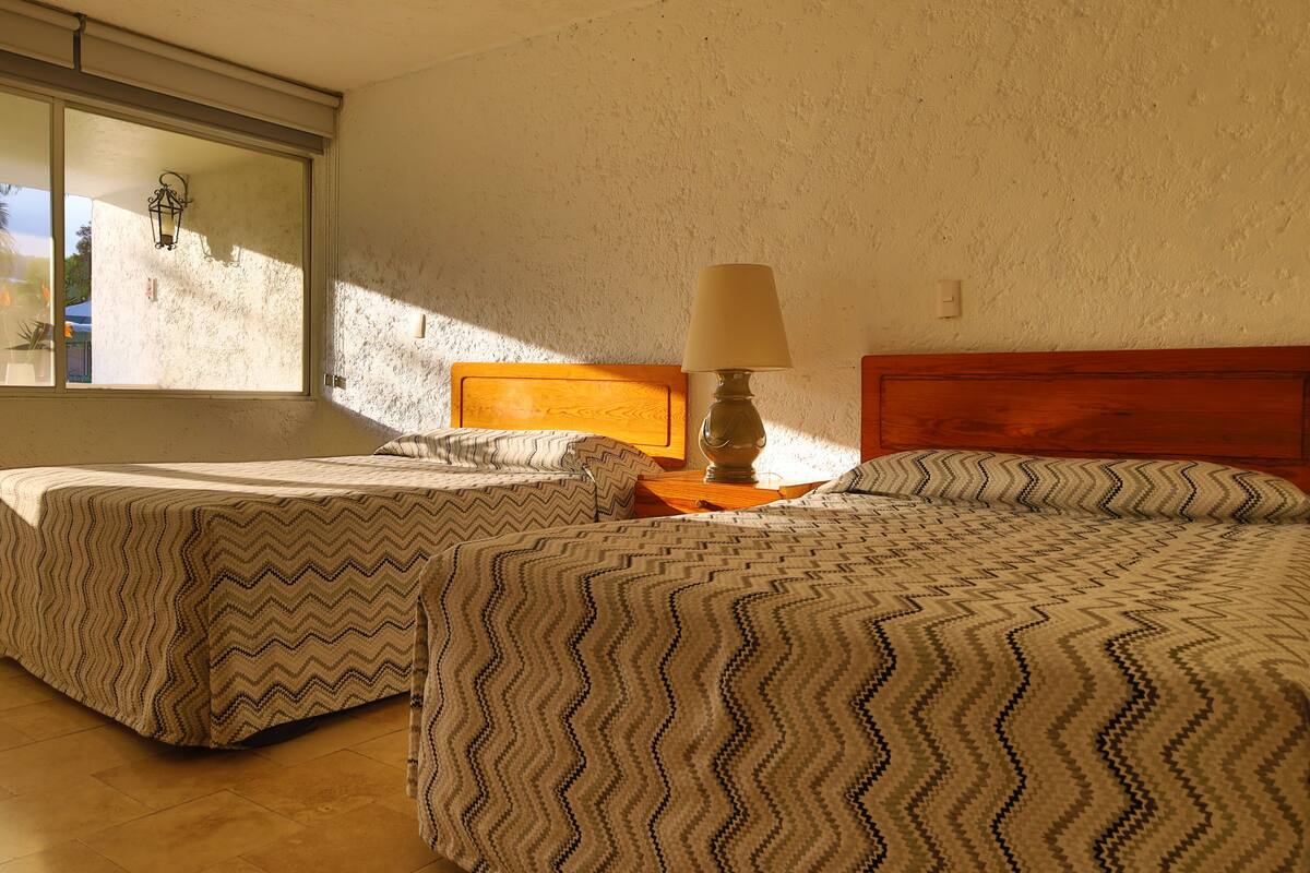 Un dormitorio con dos camas, cabeceros de madera, una mesita de noche con lámpara y una ventana con vistas al exterior.