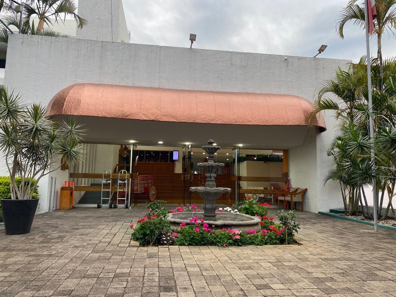 A hotel entrance with a covered patio, a central fountain, and potted plants.