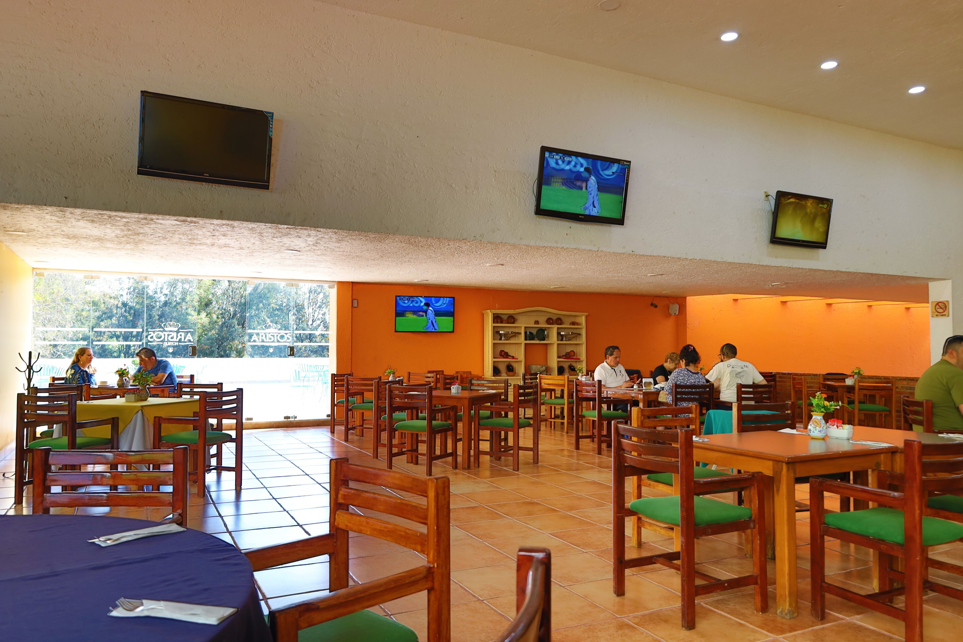 A restaurant with multiple tables and chairs, some with green cushions. There are two televisions mounted on the wall displaying sports.