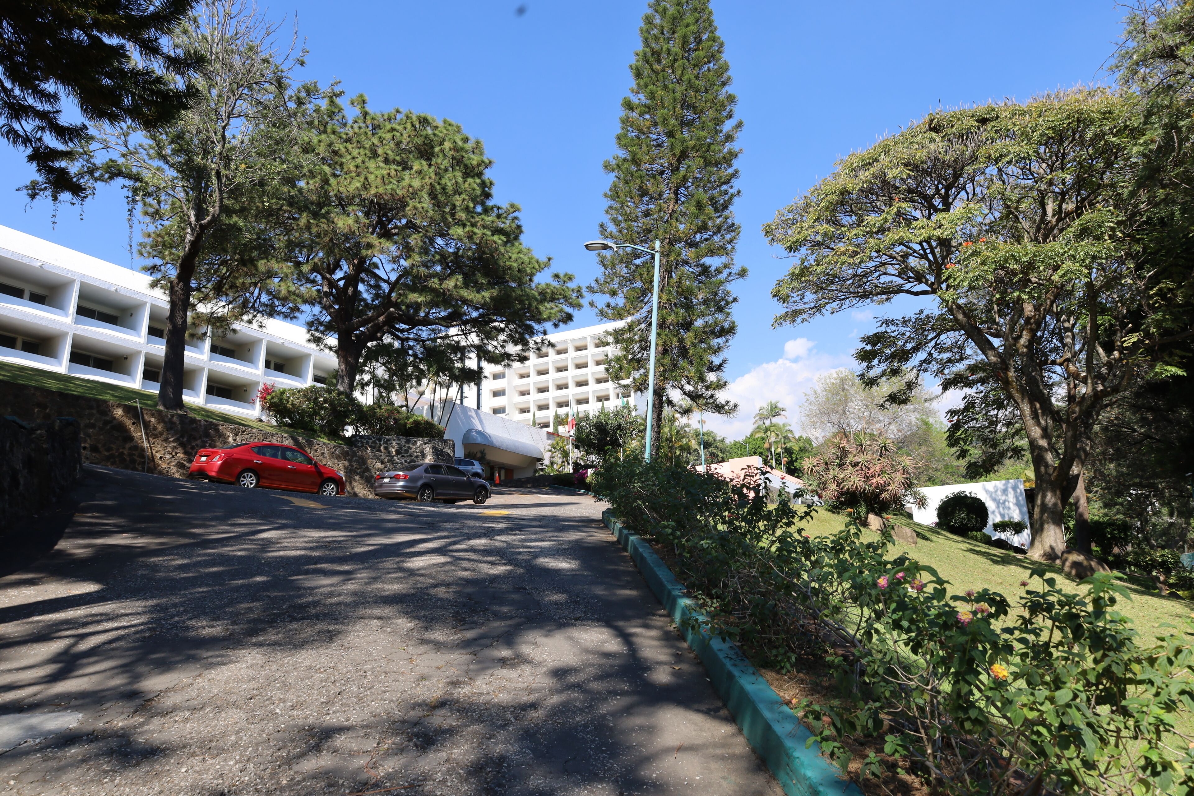 Un espace de stationnement avec une voiture rouge, un bâtiment blanc à plusieurs étages et de la verdure environnante.