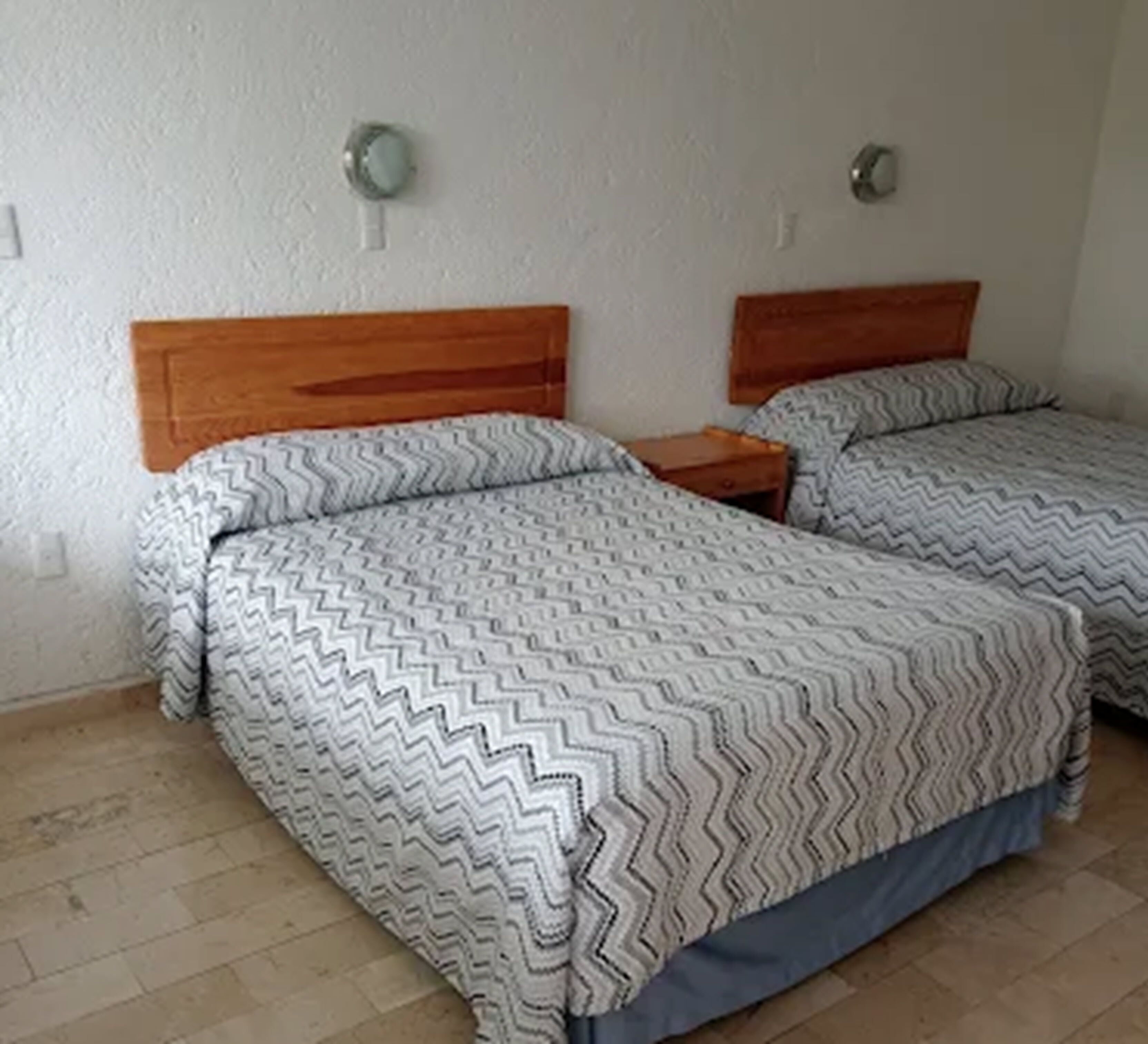 Una habitación con dos camas, cada una con cabecera de madera y colcha estampada. Hay mesitas de noche con lámparas y una pared blanca lisa.