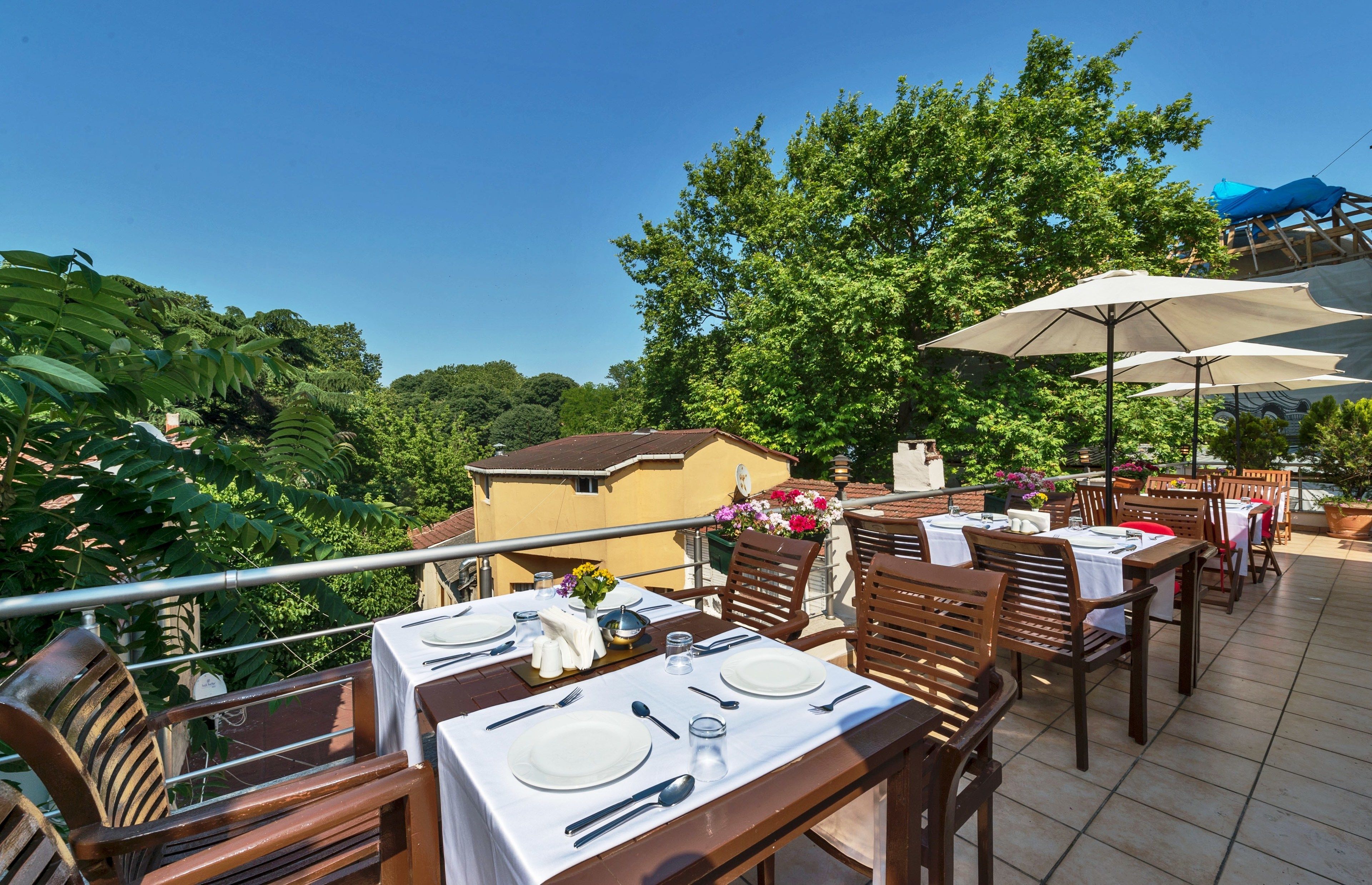 Un área de comedor al aire libre con mesas dispuestas para dos, rodeada de follaje y árboles.