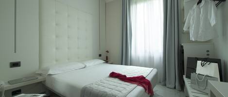 A modern hotel room with a large bed, a bedside table with a lamp, a small desk with a chair, and a window with curtains.