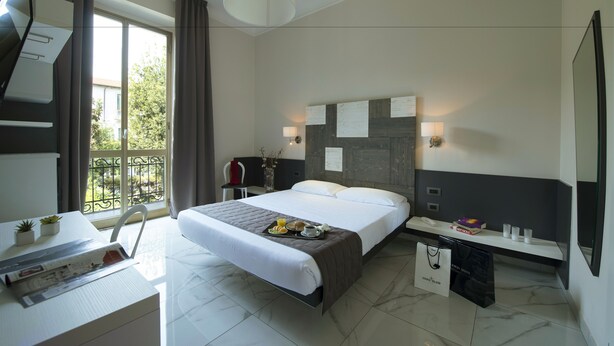 A modern hotel room with a large bed, a desk with a chair, a small table with plants, a bedside table with a lamp, and a view of greenery through the window. - Hotel San Francisco (Milan)