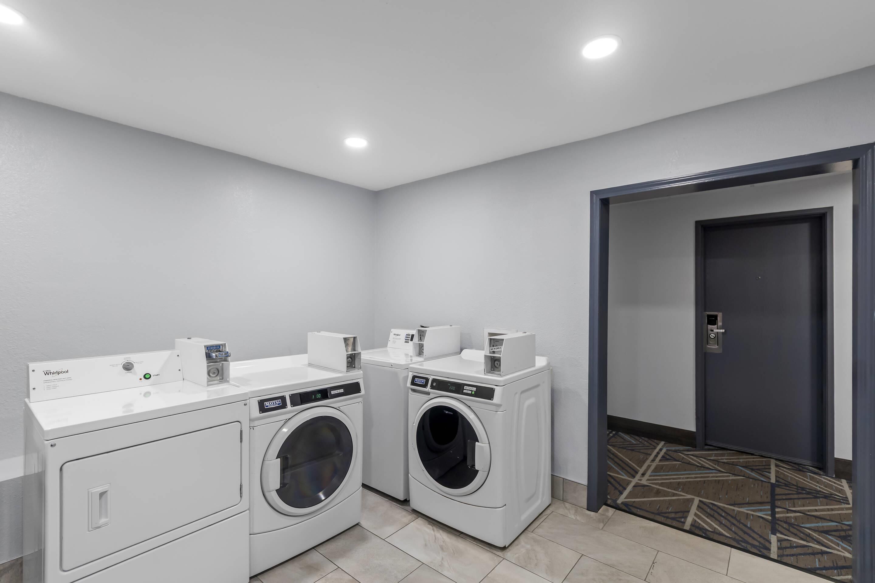 A laundry room with two front-loading washers and dryers, a control panel, and a door leading to another room.