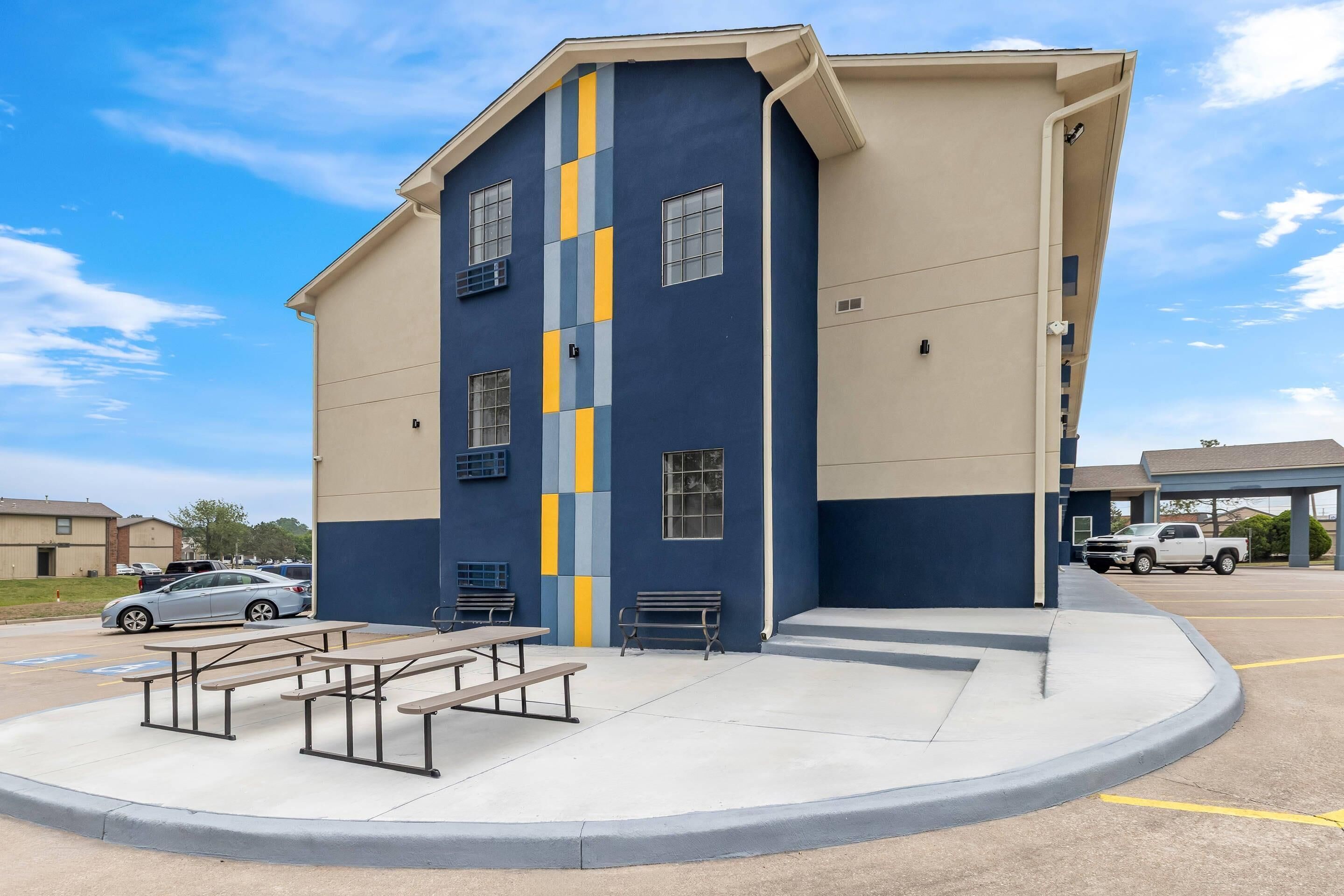 A multi-story building with a blue and beige facade, outdoor seating area, and a parking lot.