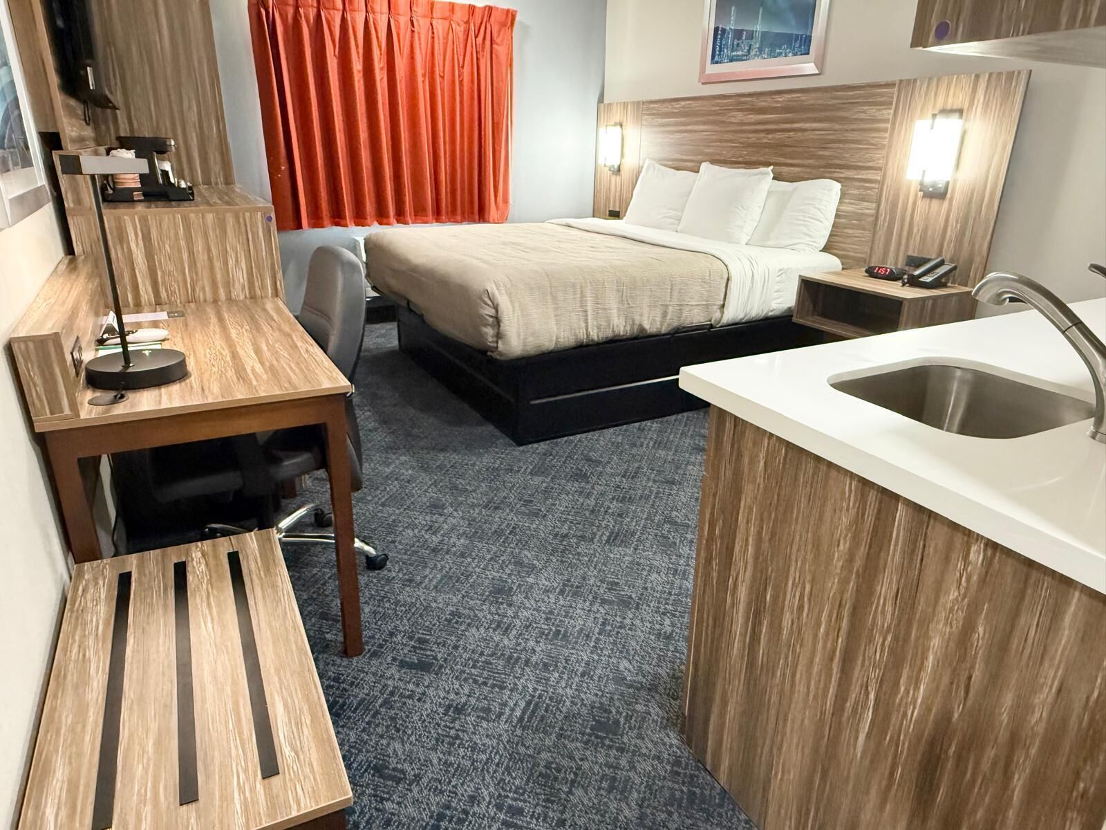 Una habitación de hotel compacta con cama, escritorio, silla, kitchenette pequeña y baño.