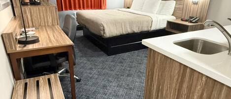 Una habitación de hotel compacta con cama, escritorio, silla, kitchenette pequeña y baño.