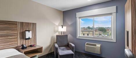 Una habitación de hotel con cama, una silla, escritorio con teléfono y lámpara, ventana con vistas y calentador de pared.