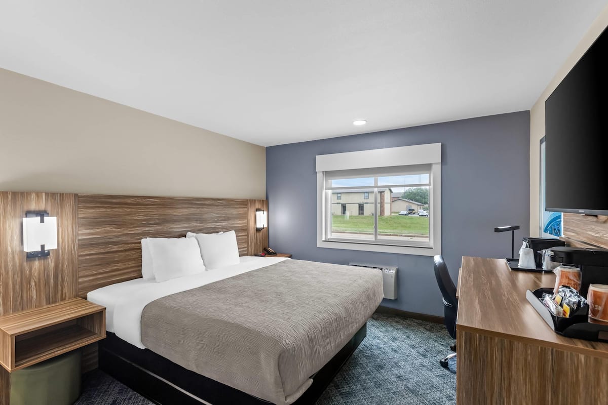 A hotel room with a bed, bedside table, desk, chair, and a window with a view of a building and greenery.