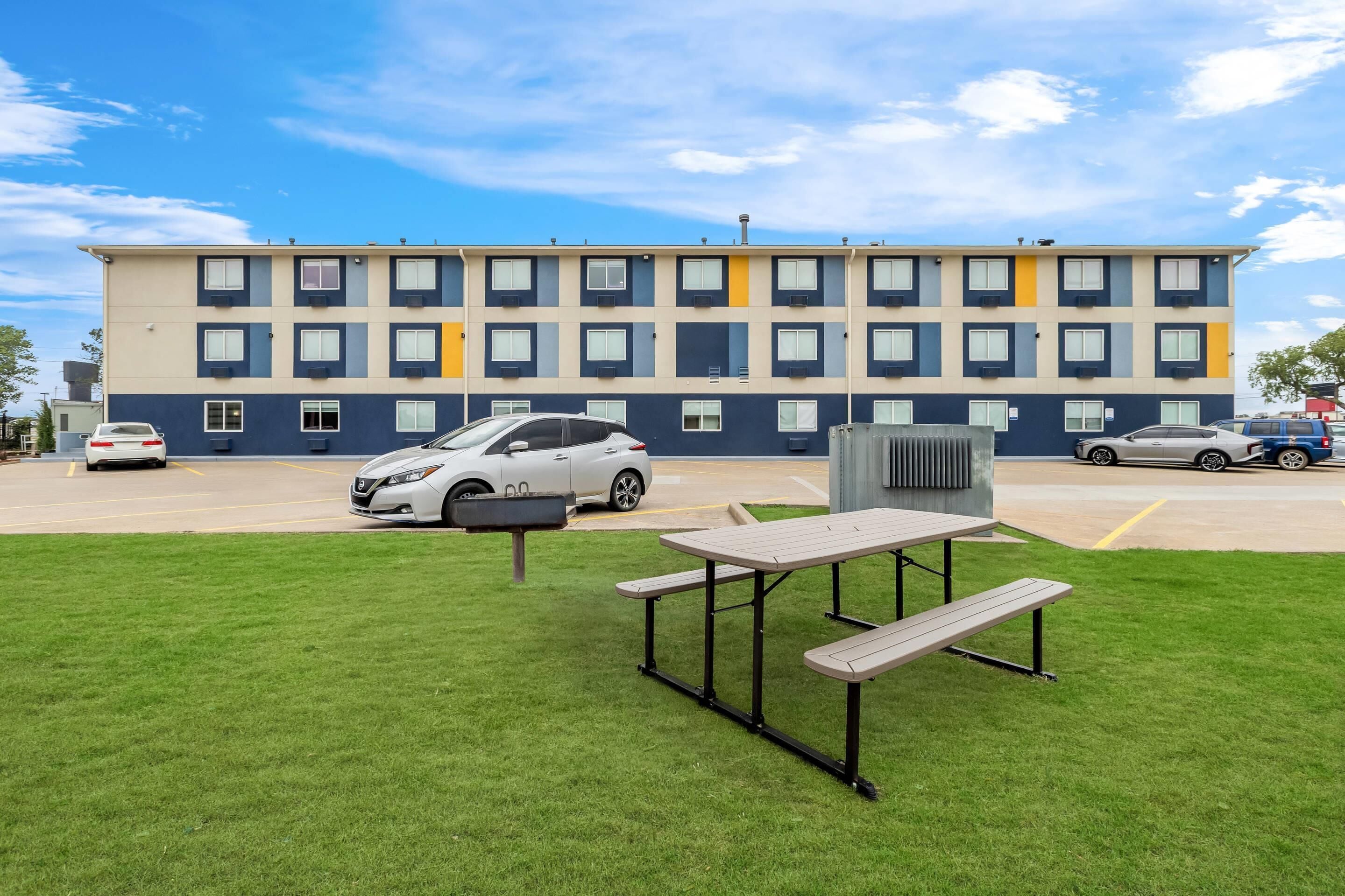 Un edificio de varios pisos con estacionamiento, una mesa de picnic y un automóvil estacionado al frente.