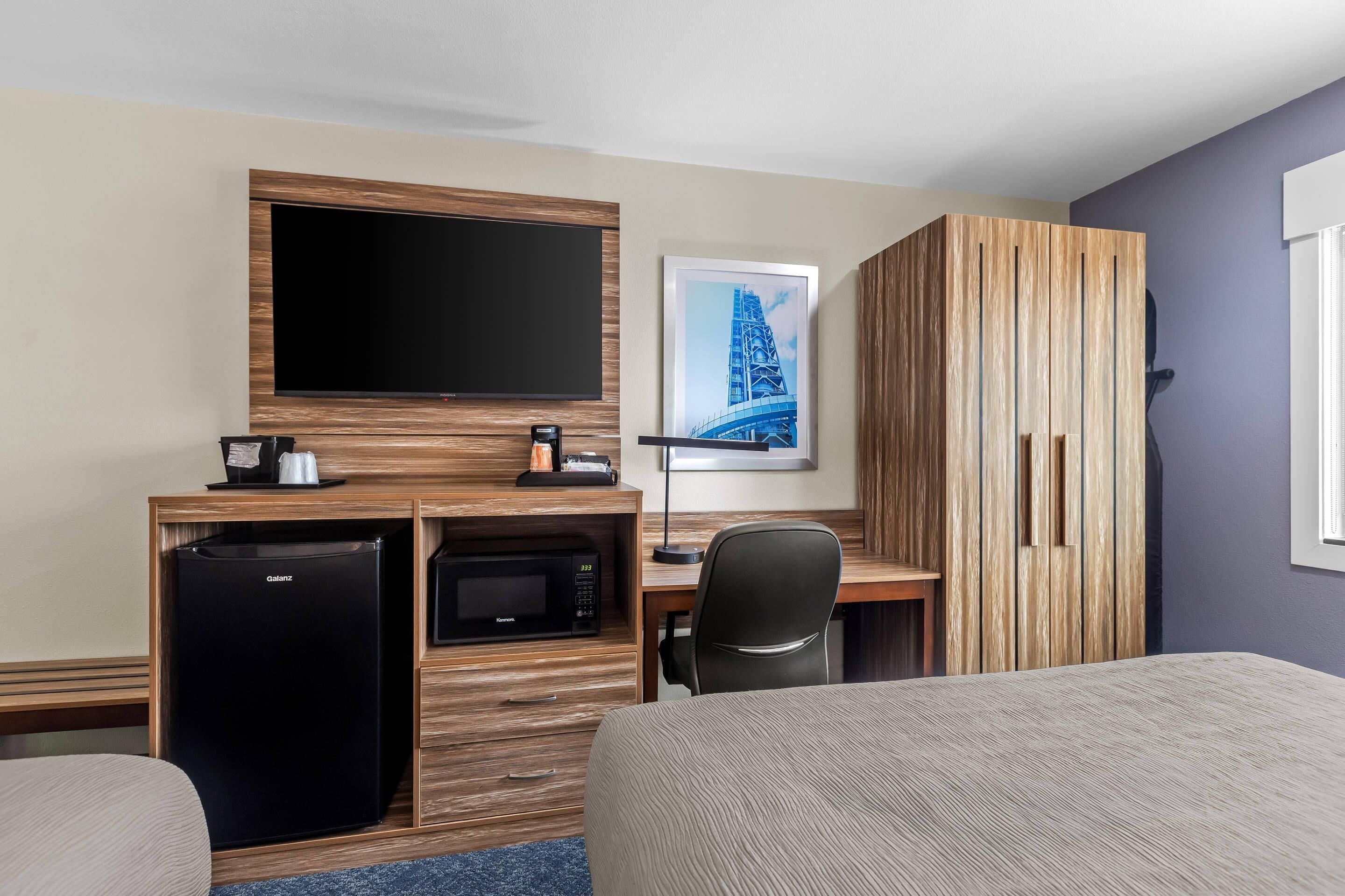 A hotel room with a bed, a desk with a flat-screen TV, a microwave, a refrigerator, a chair, and a wardrobe.