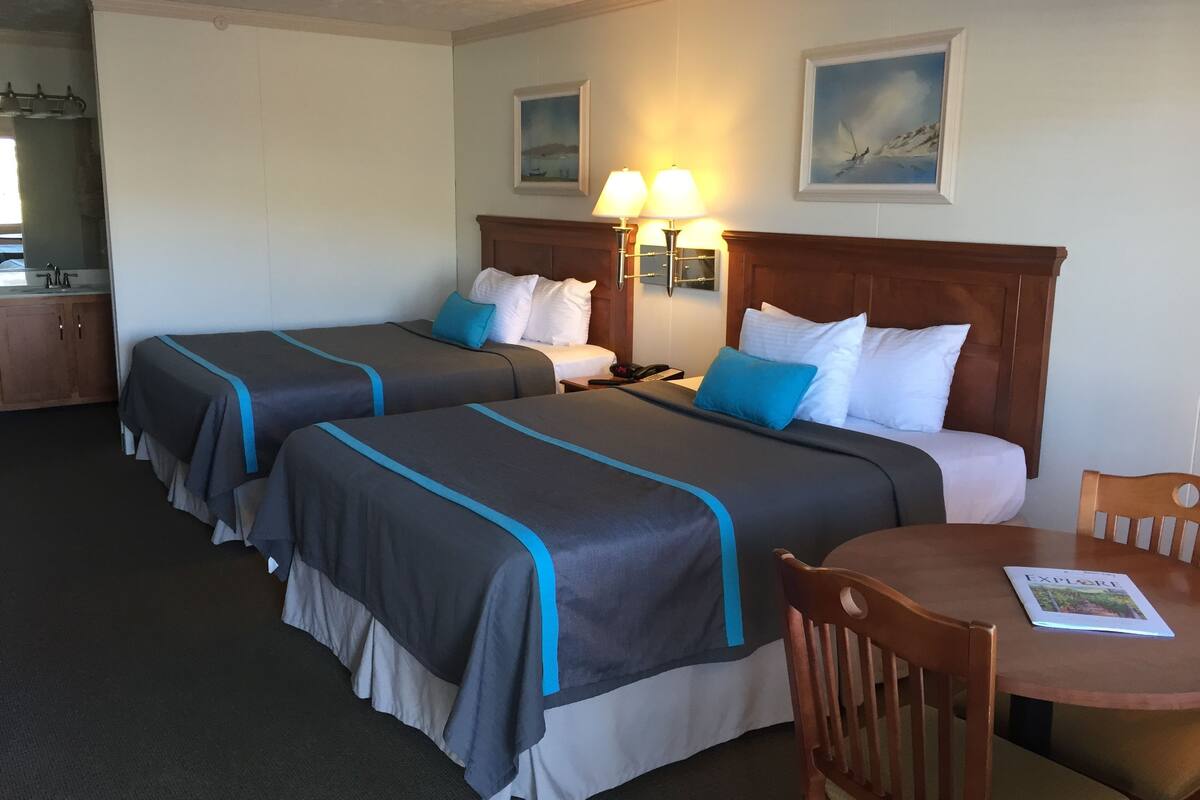 Ein Hotelzimmer mit zwei Betten, einem hölzernen Kopfteil, einem kleinen Tisch und einem Stuhl. An der Wand befinden sich zwei Wandleuchten und zwei gerahmte Bilder.