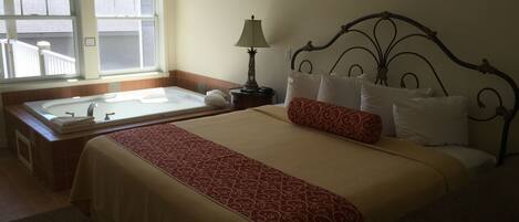 Habitación de hotel con una cama grande, ventana con vistas, baño con bañera y mesita de noche con lámpara.