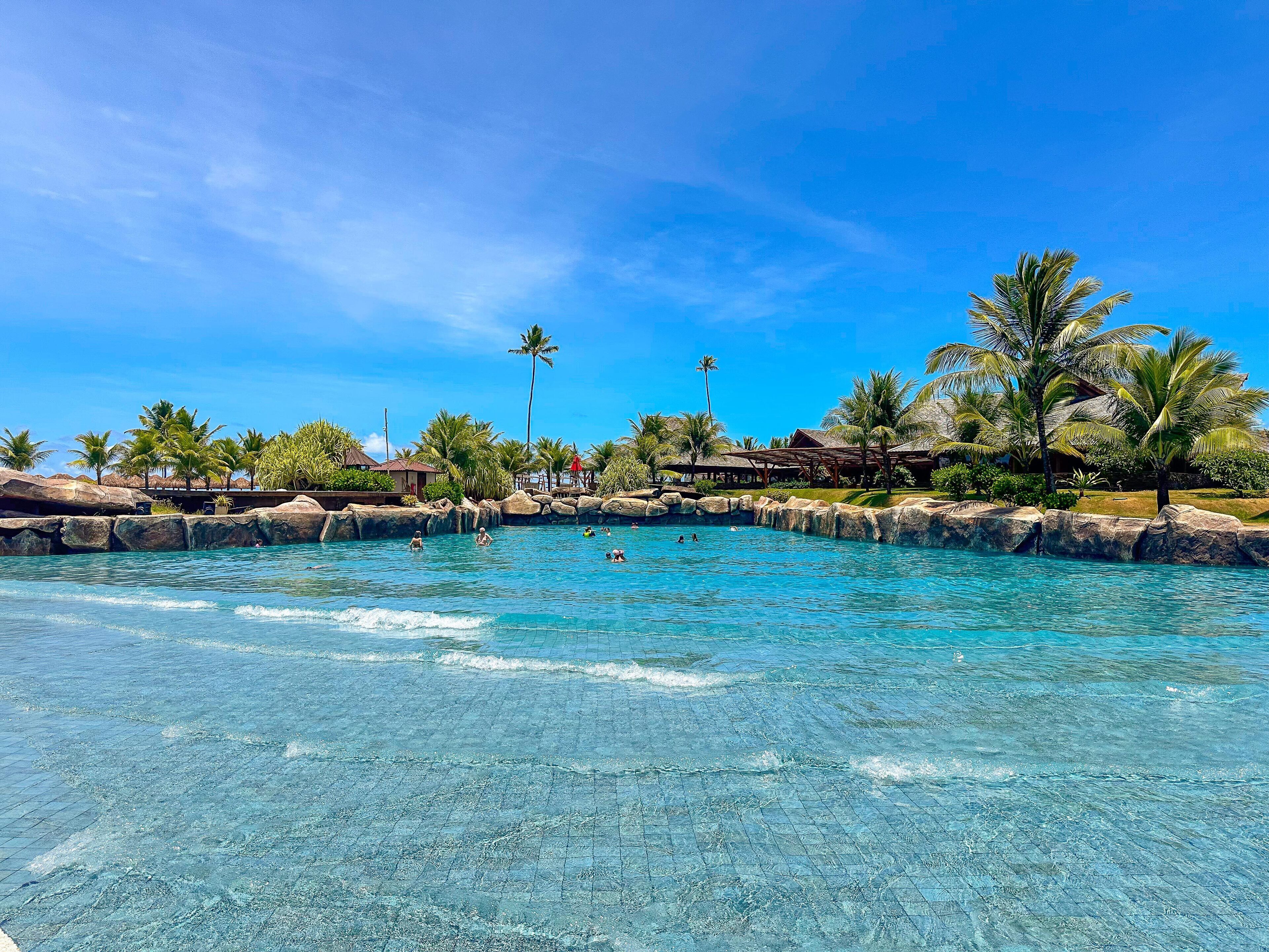 A clear pool with rocks and palm trees, surrounded by buildings.