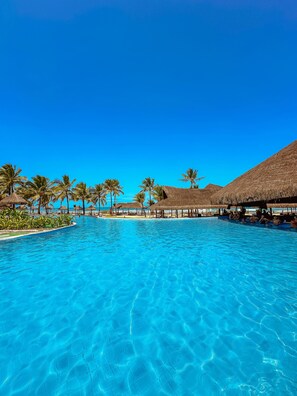 A clear blue pool with thatched-roof structures and palm trees. - Enotel Porto de Galinhas - All inclusive (Ipojuca)