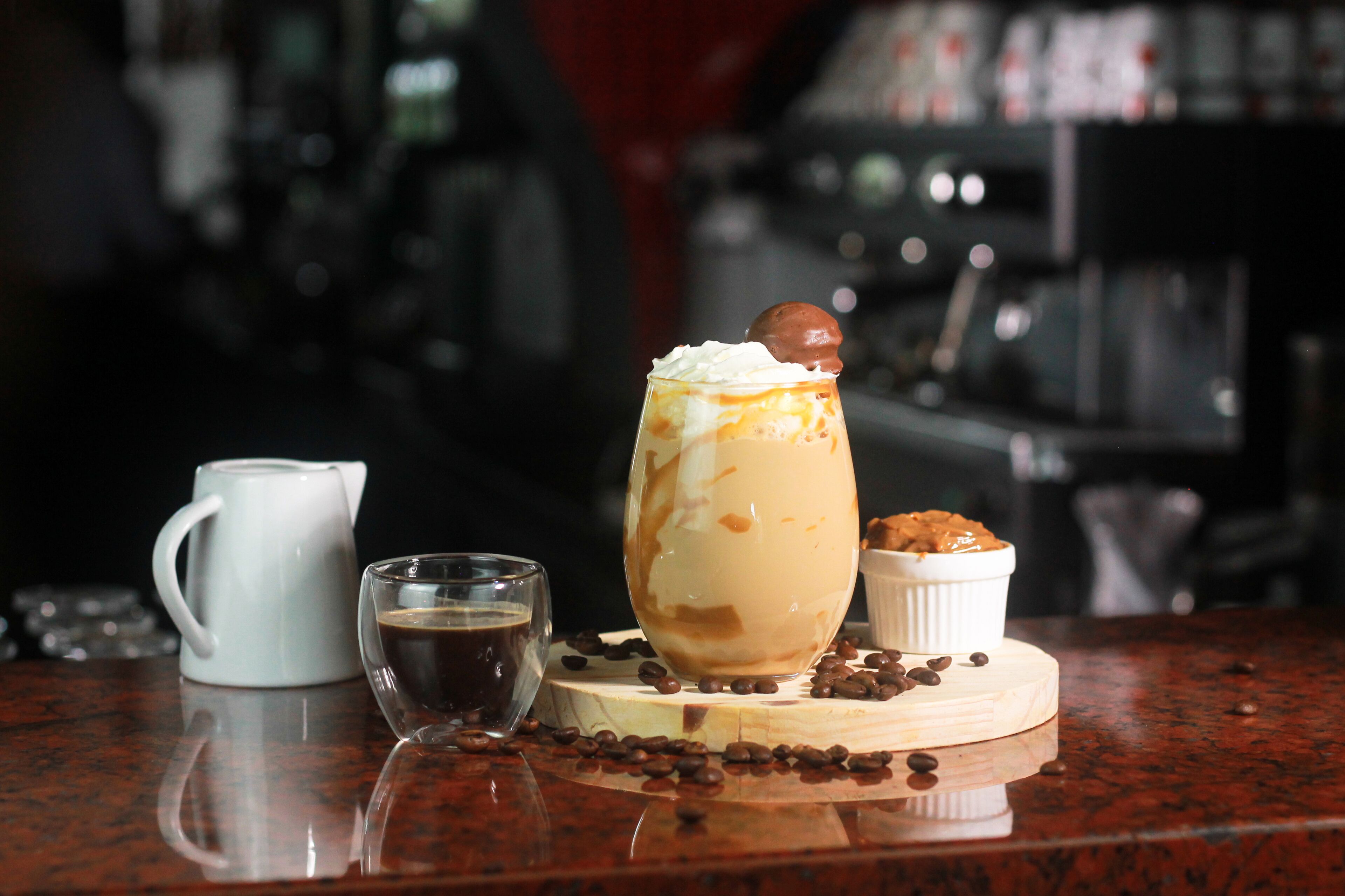 A dessert with whipped cream and a chocolate ball on top, served in a tall glass, with a small cup of coffee and a pitcher of coffee on a wooden board, surrounded by coffee beans.