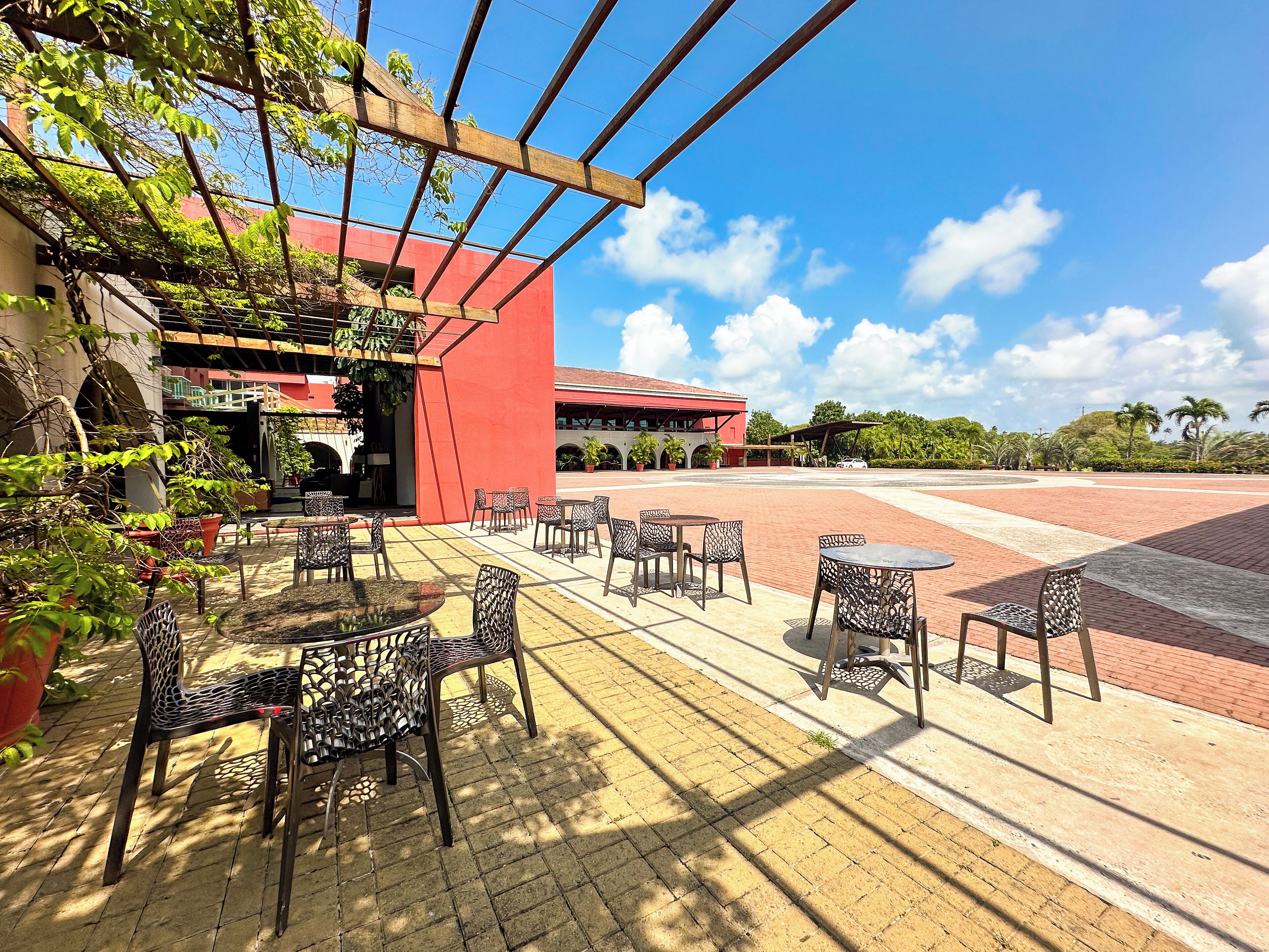 Un patio al aire libre con mesas y sillas, un edificio rojo al fondo y un cielo azul despejado.