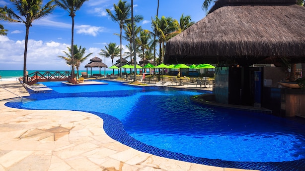 Zona de piscina en un resort, con vista despejada al océano, rodeada de palmeras y estructuras de techo de paja.