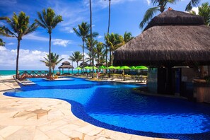 Zona de piscina en un resort, con vista despejada al océano, rodeada de palmeras y estructuras de techo de paja.