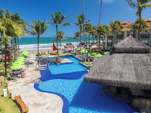 A resort pool area with a thatched-roof structure, palm trees, and a view of the ocean. - Solar Porto de Galinhas (Ipojuca)