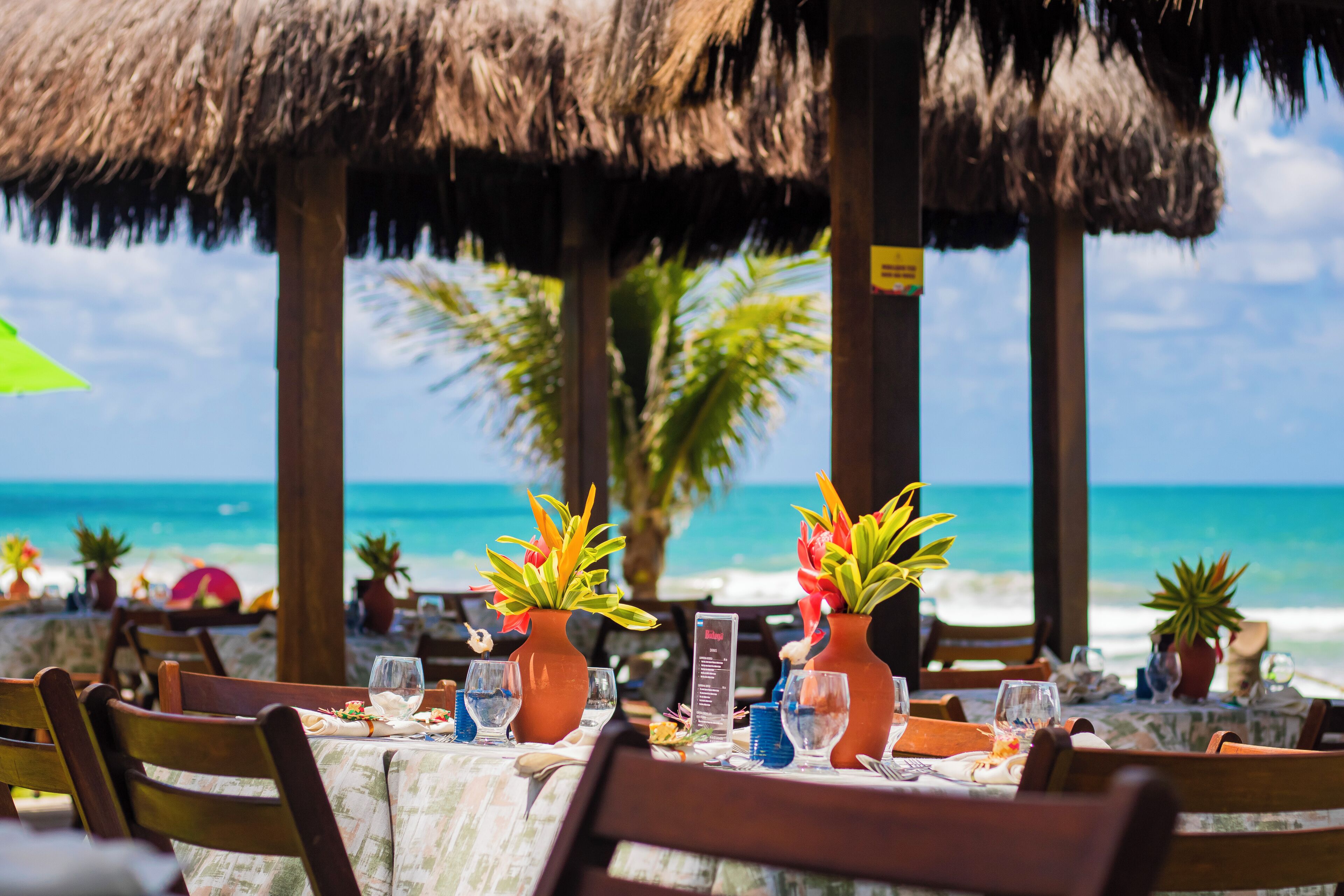 Un restaurant situé en bord de mer, avec des tables dressées pour le dîner, des parasols en chaume et une vue sur l’océan.