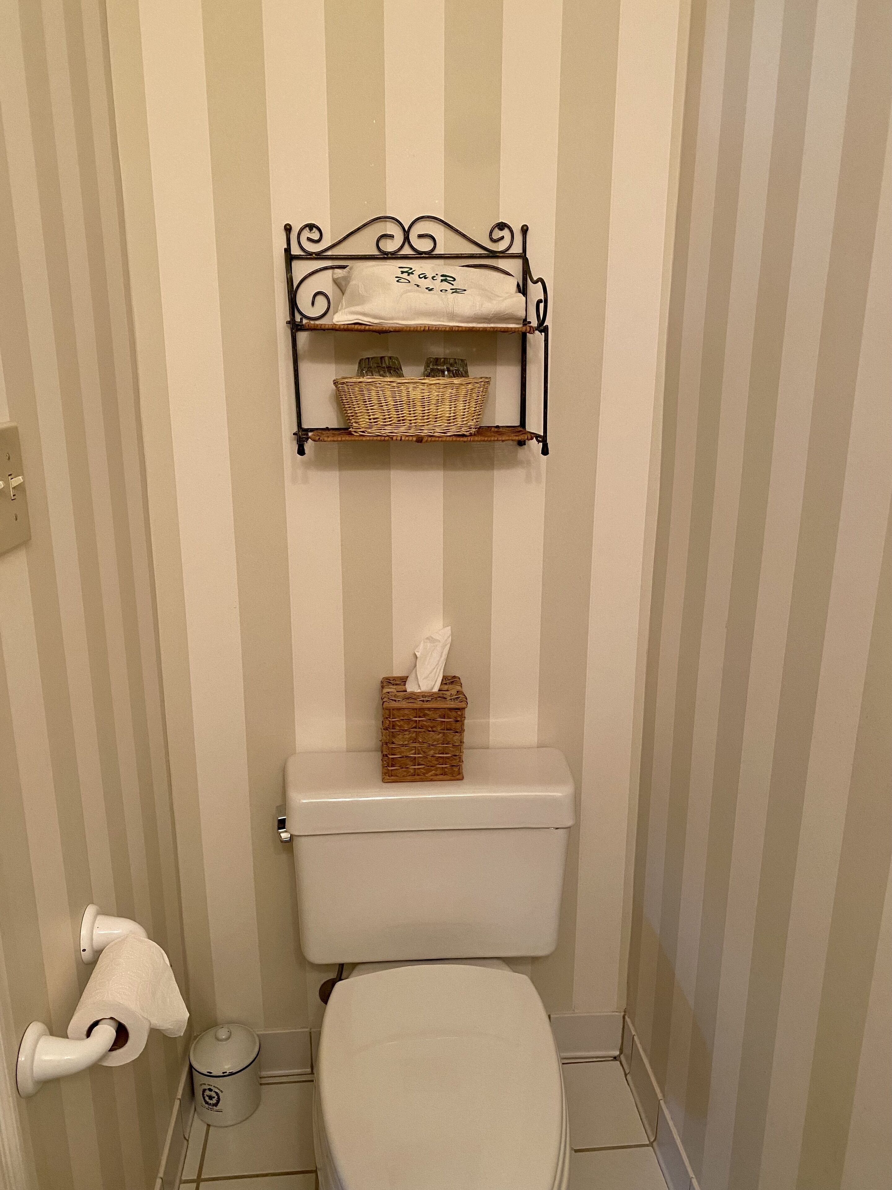 A bathroom with a toilet, a tissue holder, and a basket of toiletries on a shelf above the toilet.