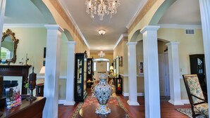 Un amplio pasillo con un gran jarrón decorativo en el centro, columnas blancas, candelabros y piso de madera.