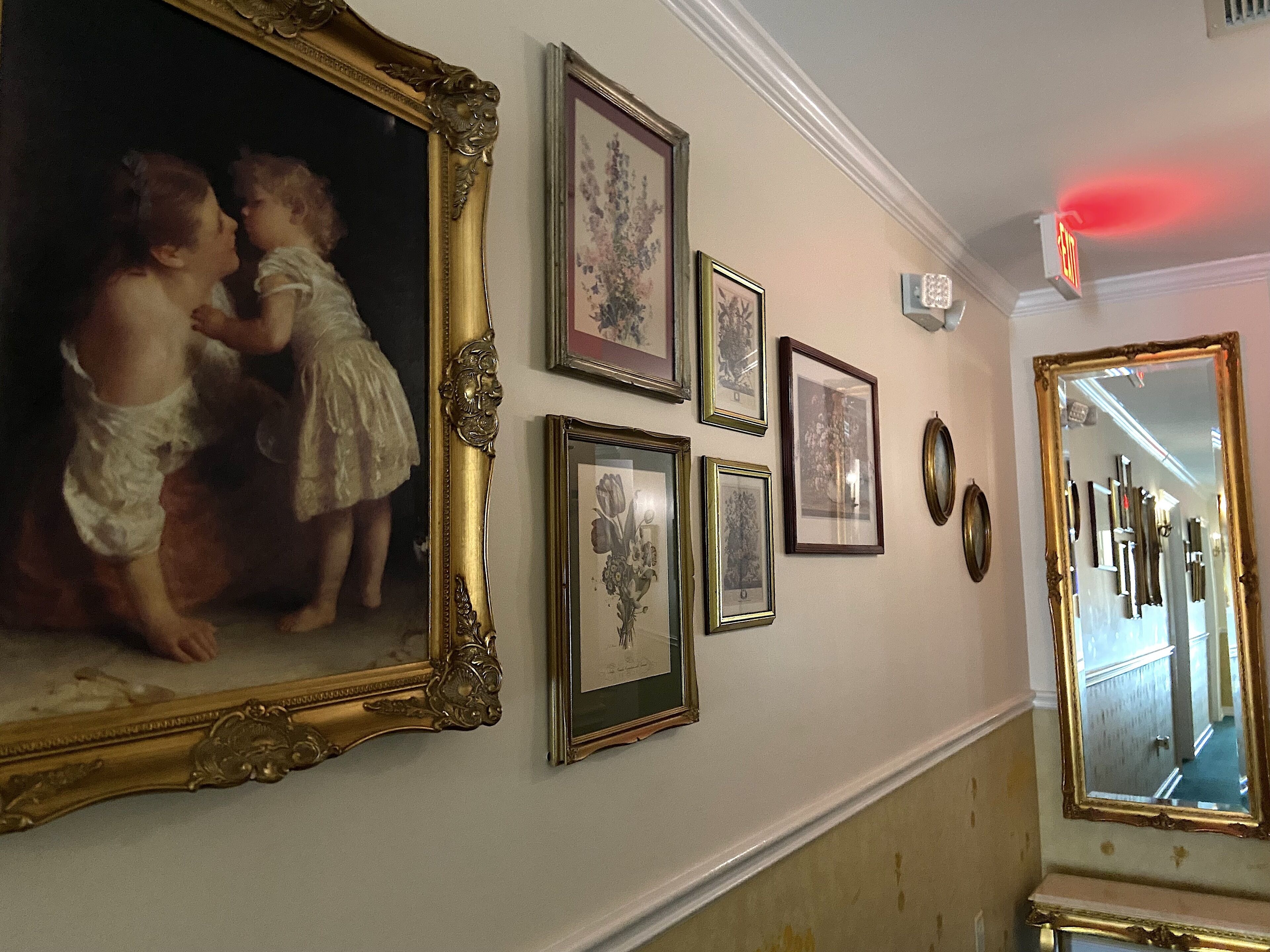 A hallway with a large ornate mirror, several framed pictures on the wall, and a red exit sign.