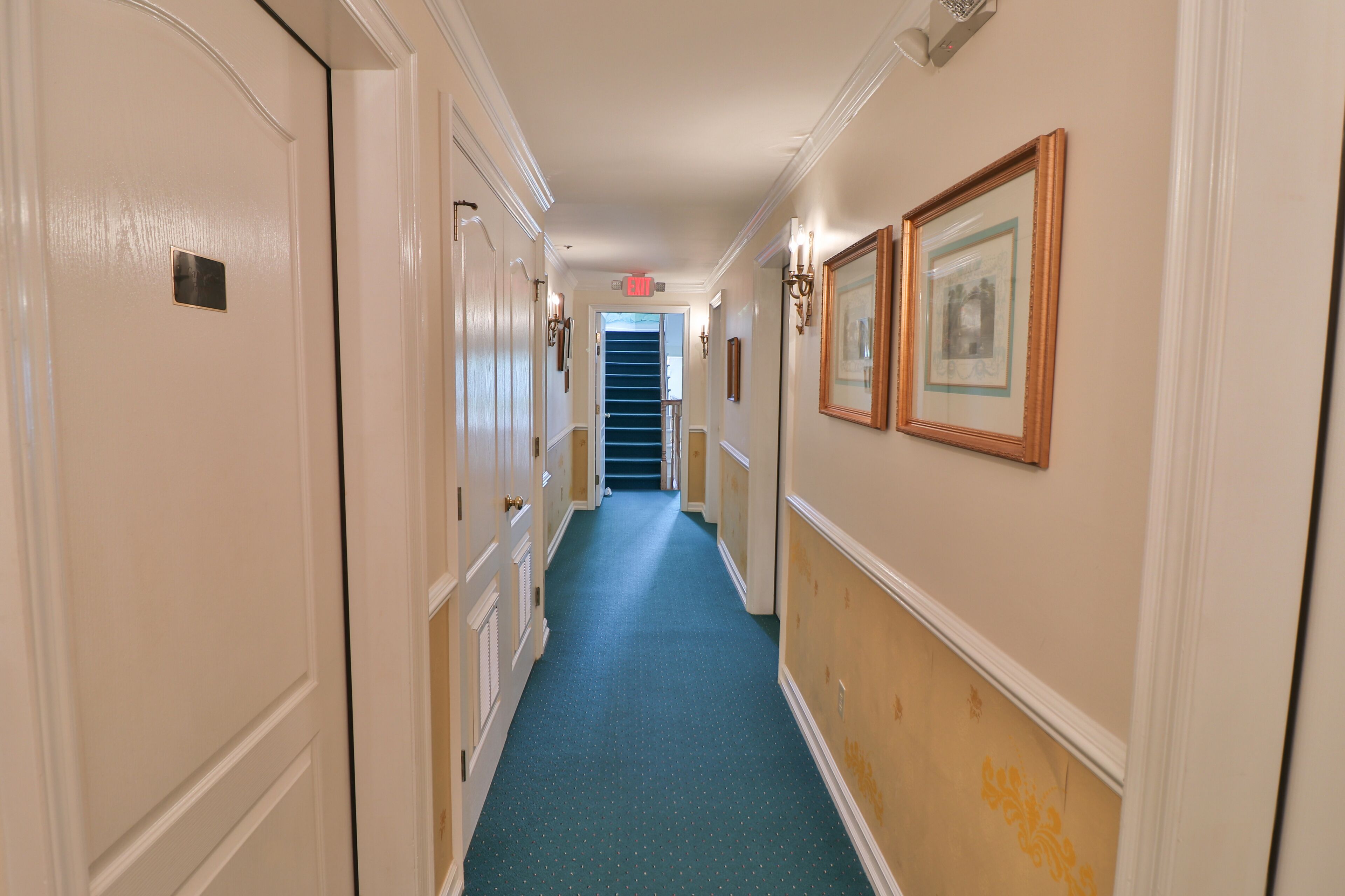 A hallway with cream-colored walls, a blue carpet, and framed pictures on the right.