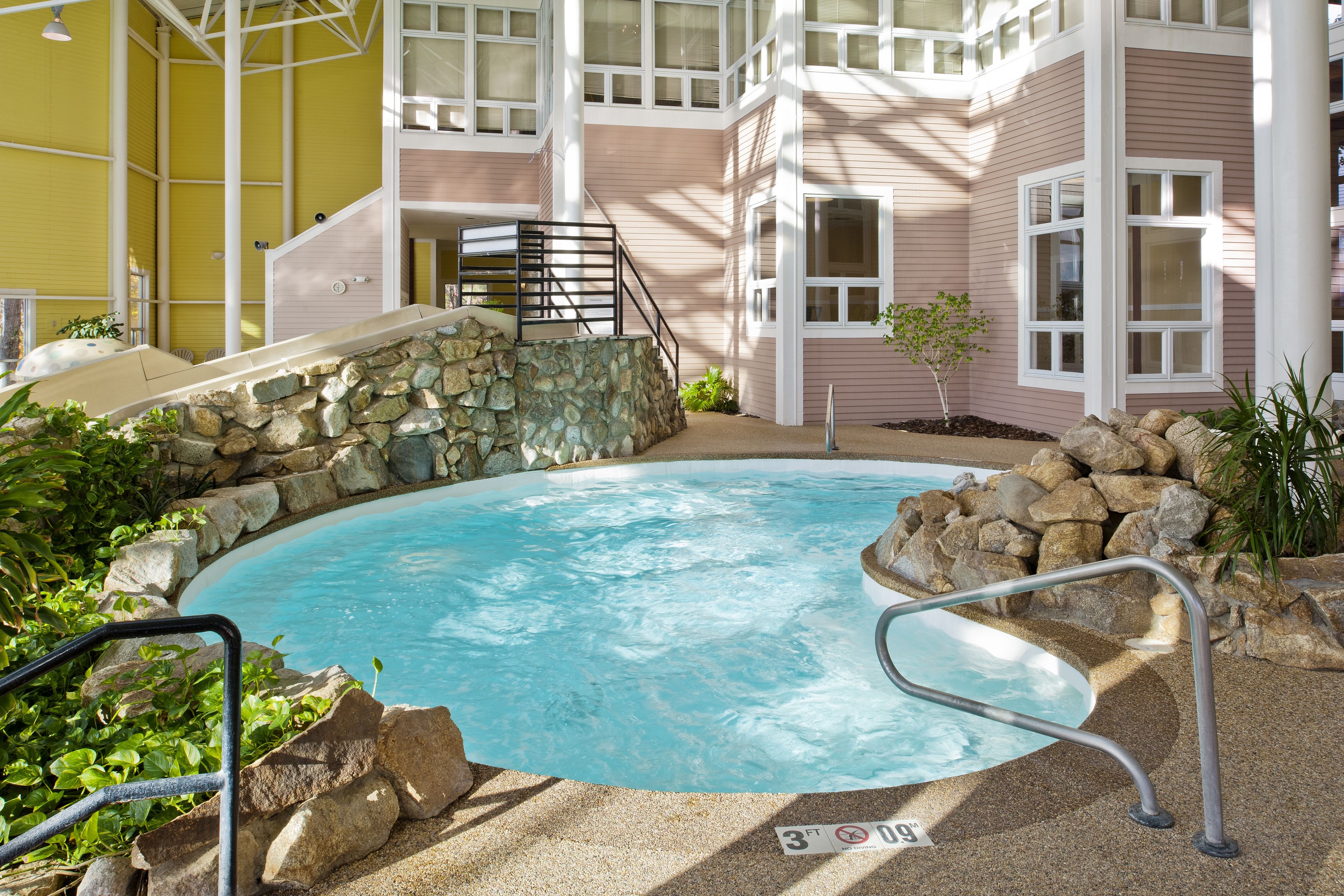 A pool area with a stone wall, metal railings, and a staircase leading to a building with large windows.