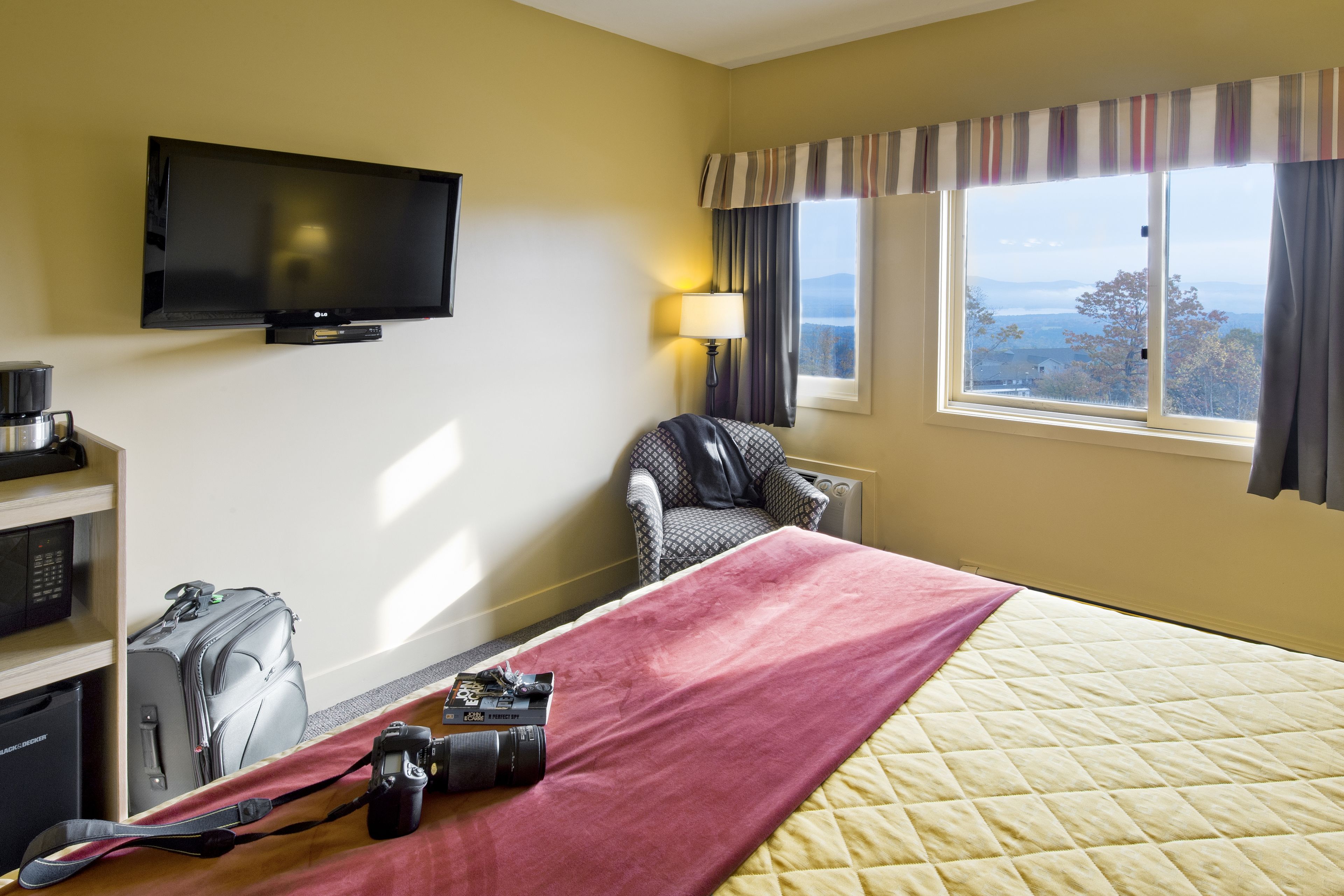 A hotel room with a bed, a television mounted on the wall, a desk with a lamp, a chair, and a view of the outdoors through a window.