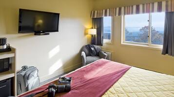 A hotel room with a bed, a television mounted on the wall, a desk with a lamp, a chair, and a view of the outdoors through a window.