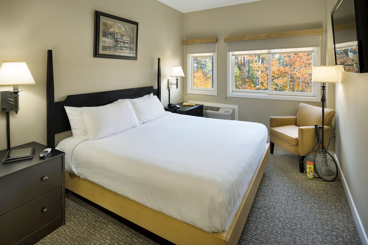A hotel room with a bed, bedside lamps, a chair, a table, and windows with autumn foliage views.