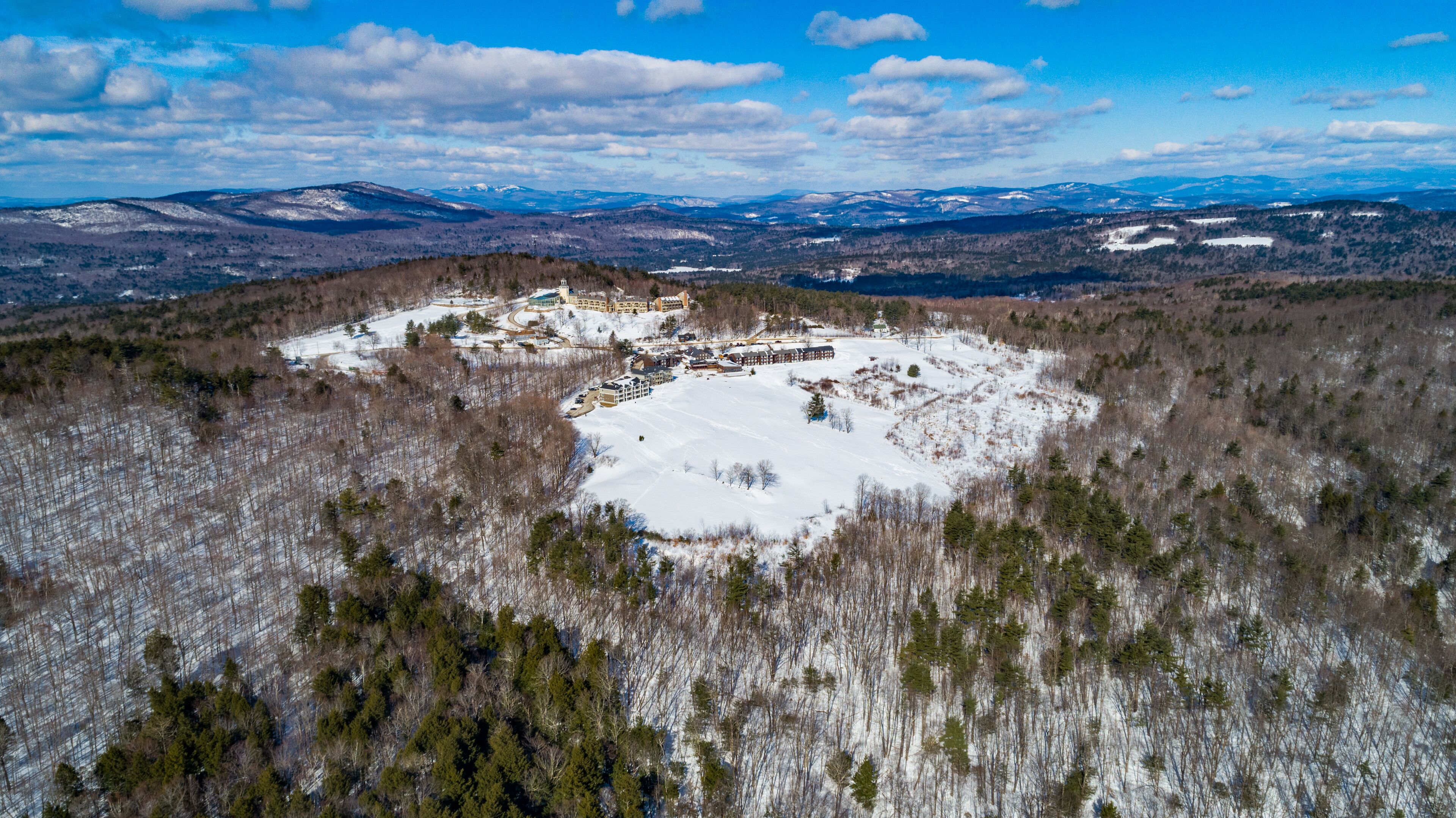 Vista aérea de un resort con varias construcciones, una amplia zona nevada y colinas boscosas circundantes.