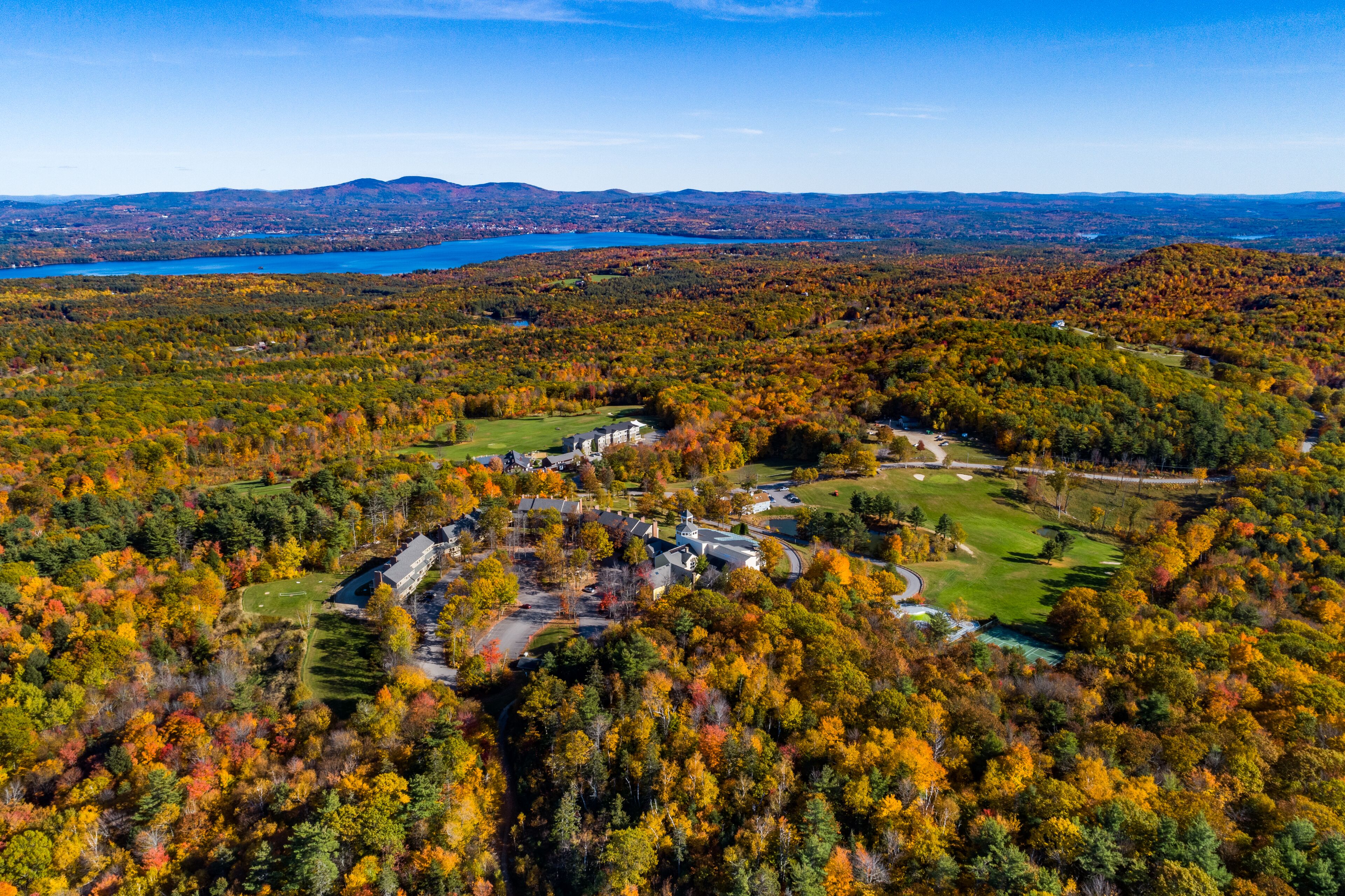 An aerial view of a resort with a golf course, surrounded by autumn-colored trees and a lake in the background.
